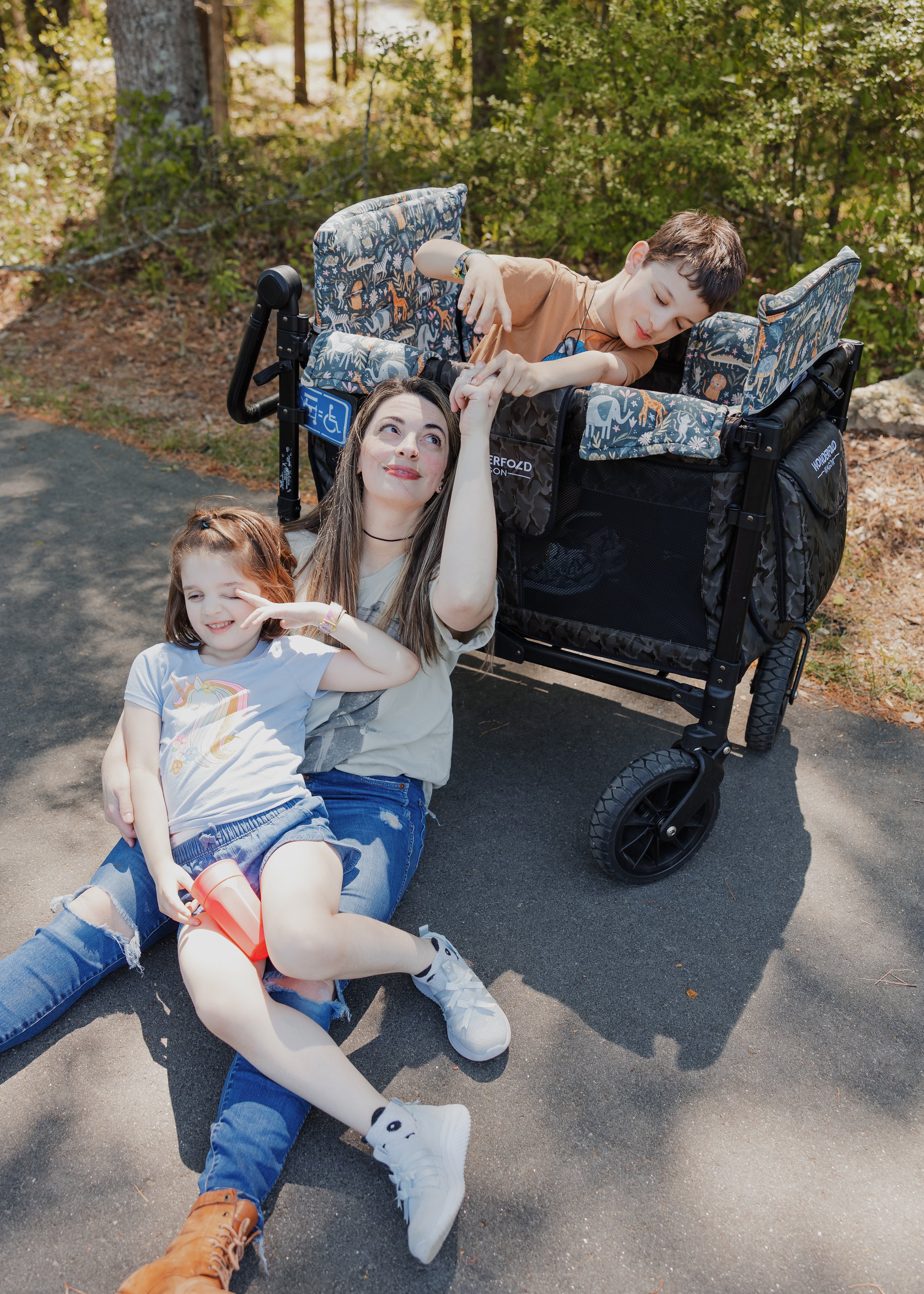 A mother sits on a shaded Wake Forest greenway with her two profoundly autistic, largely nonverbal children, who relax and play around a WonderFold wagon during a child-led family photo session.