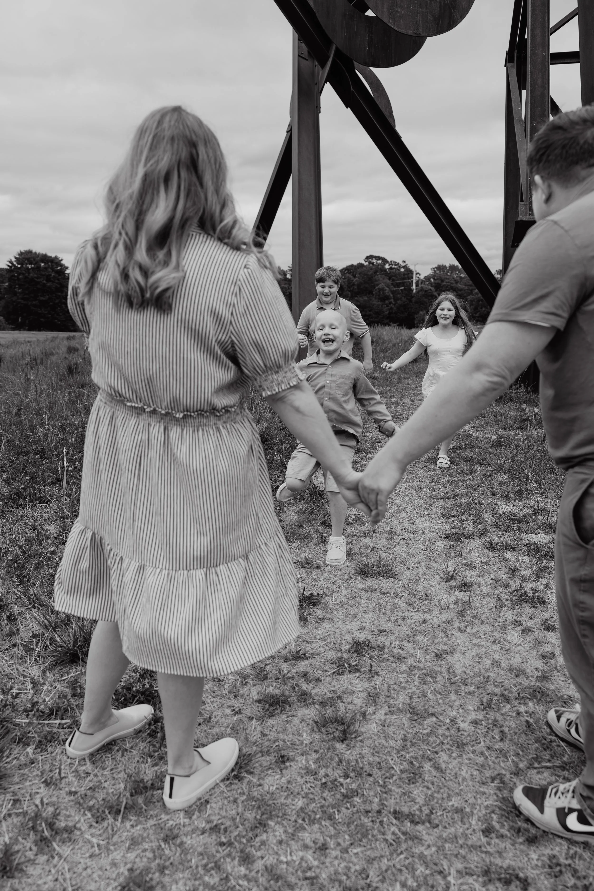 A joyful child with alopecia runs toward his parents while his siblings follow behind during a child-led family photo session at the North Carolina Museum of Art in Raleigh, NC.