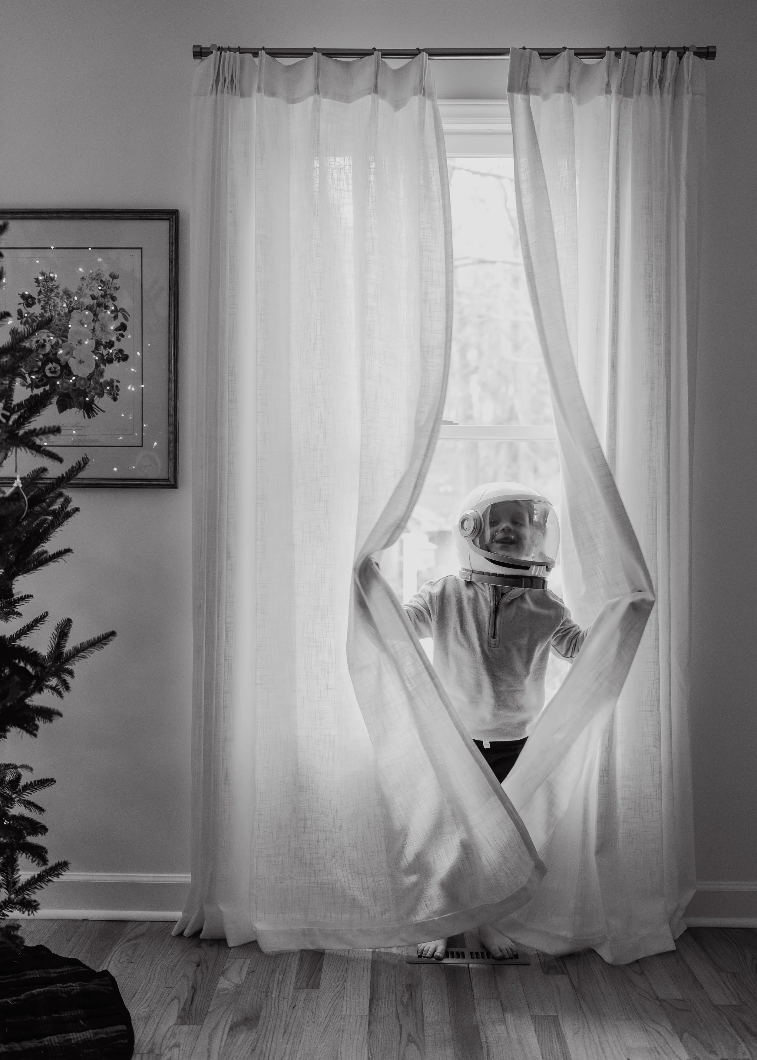 Young boy in an astronaut helmet playing behind curtains during a relaxed, full-length family photography session in Durham, North Carolina.
