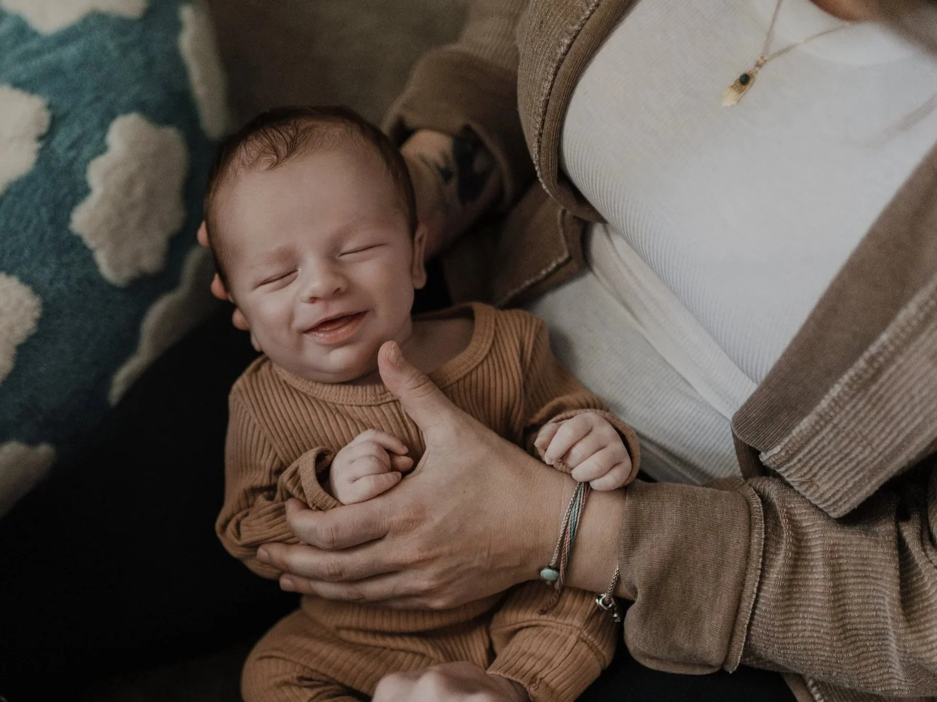 Photo of a smiling newborn baby laying in mother's lap during a baby-led photo session captured at home.