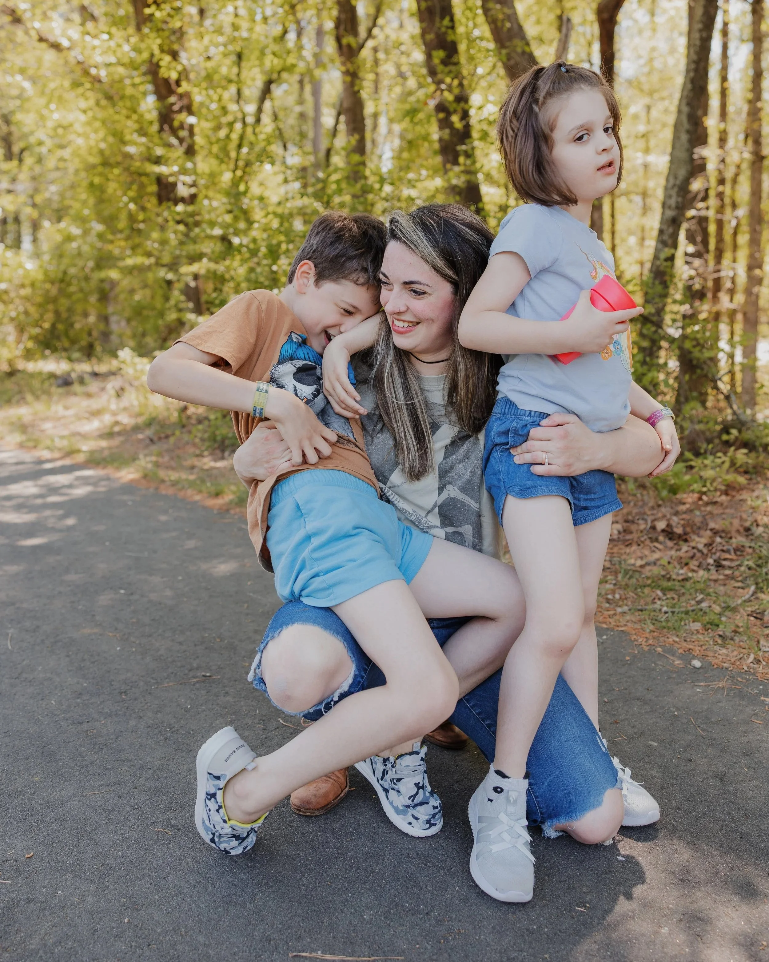 Mother kneels on a Wake Forest greenway while hugging her two profoundly autistic, largely nonverbal children during a playful family photo session.