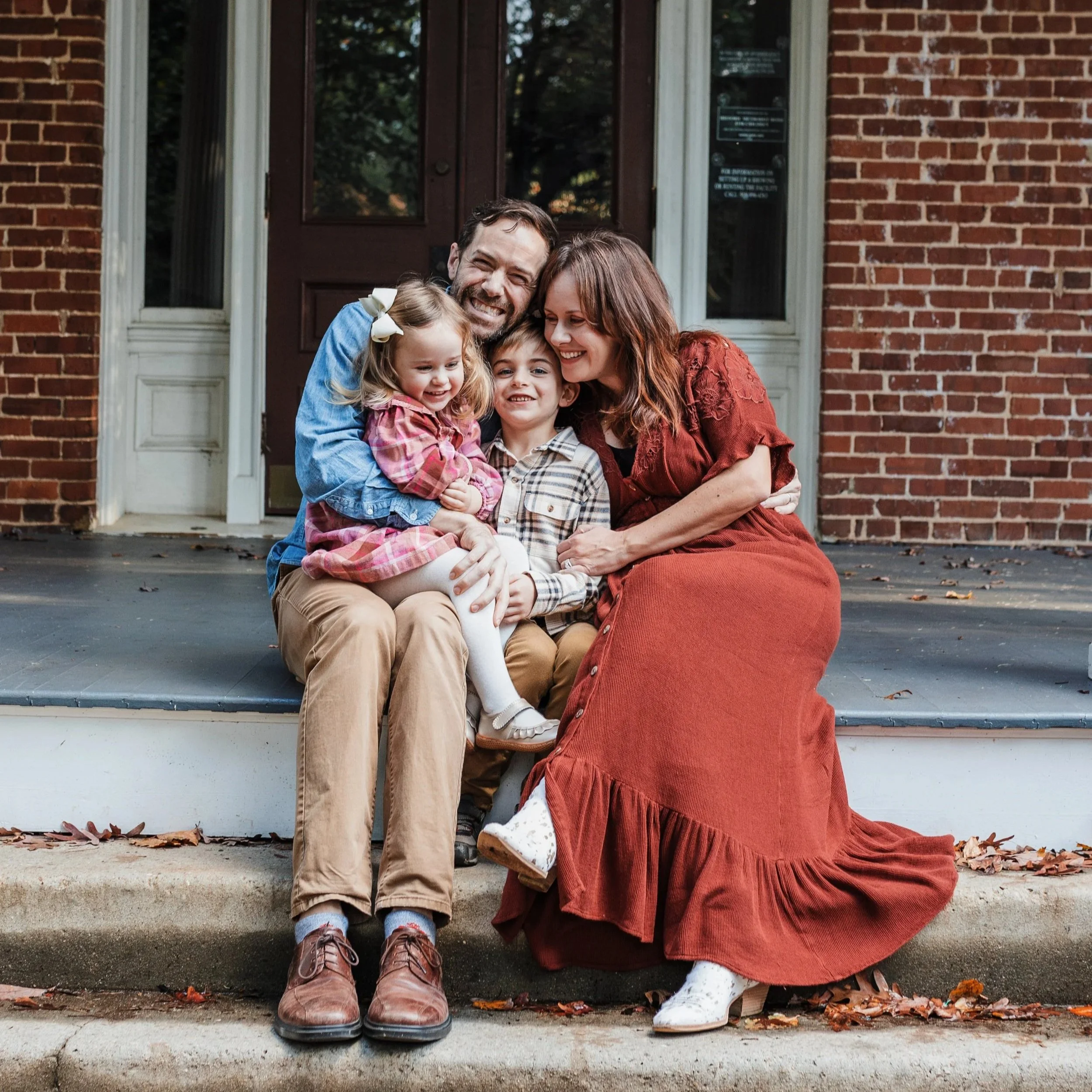 Family with a 5-year-old and toddler sit together on steps in front of a brick building during a lifestyle family photo session at Fred Fletcher Park in Raleigh.