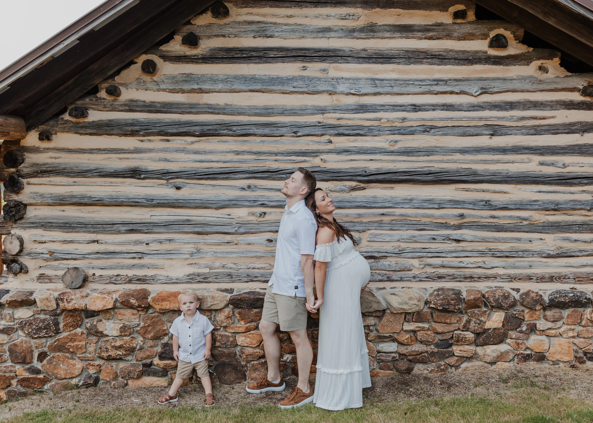 Family with pregnant mother standing against rustic wooden wall during an outdoor maternity session in Raleigh.