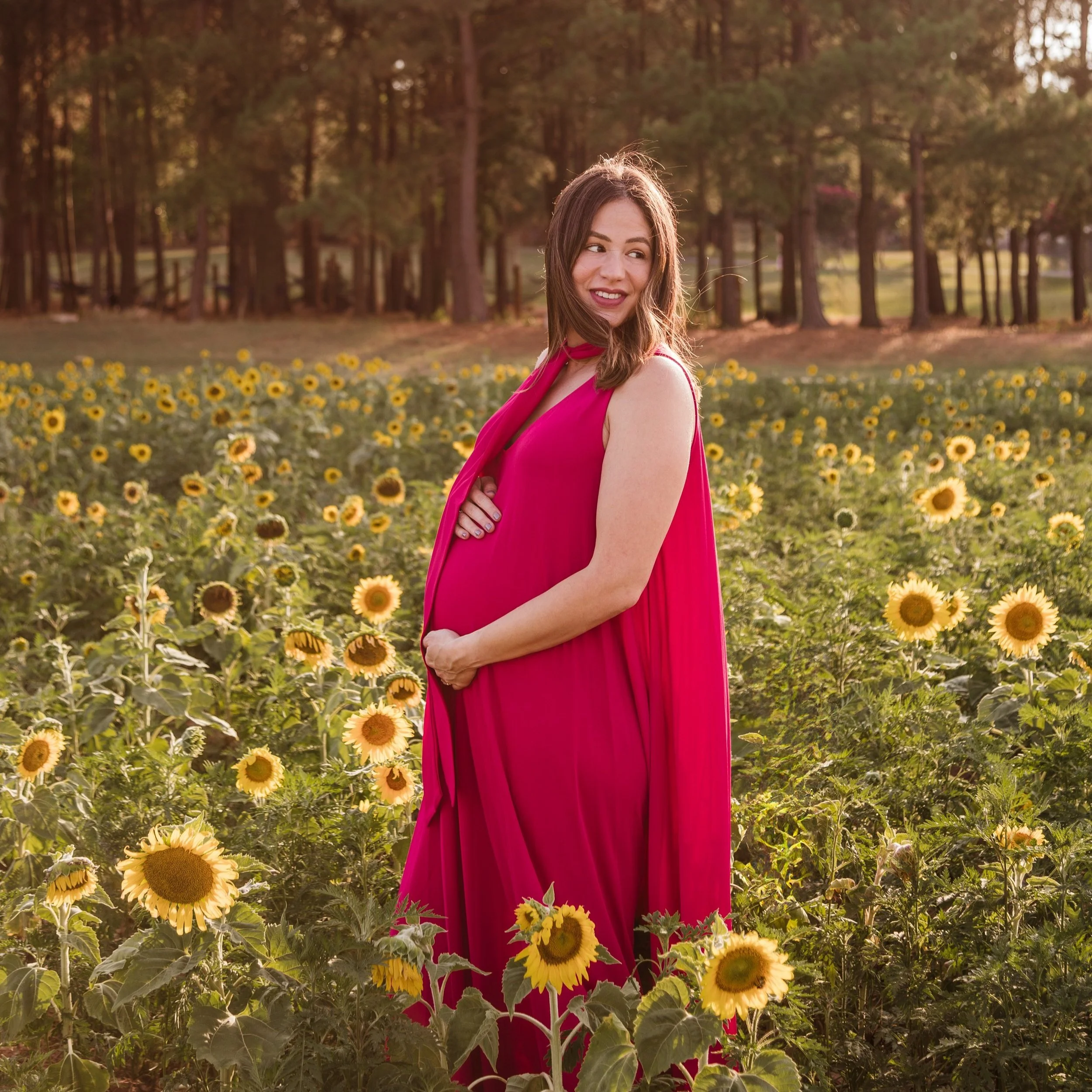 Pregnant woman in flowing red dress standing in a sunflower field during golden hour, captured by a Raleigh maternity photographer