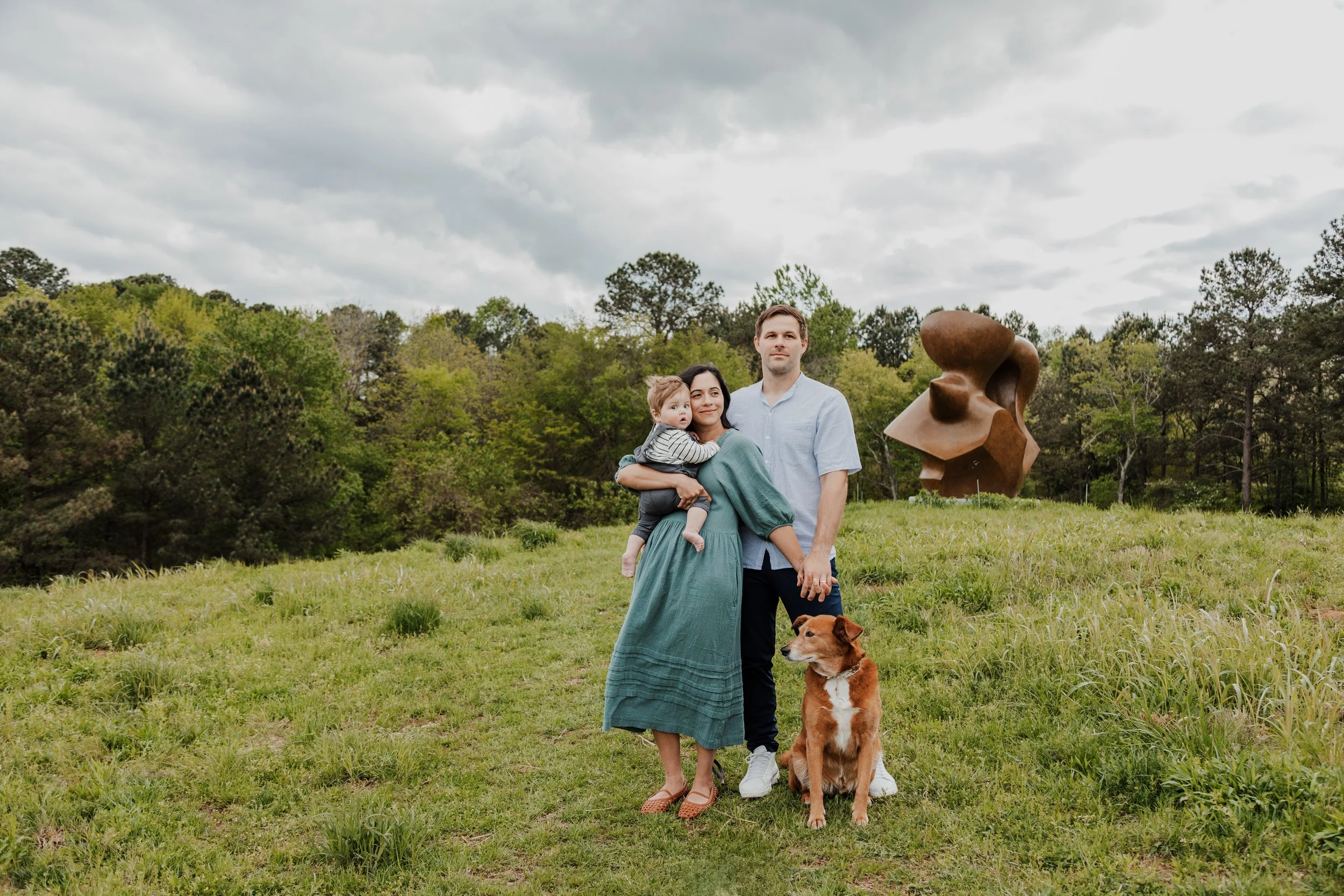 Husband and wife pose with baby and dog on grassy hill with a sculpture behind them at the Ann and Jim Goodnight Park in Raleigh, NC.