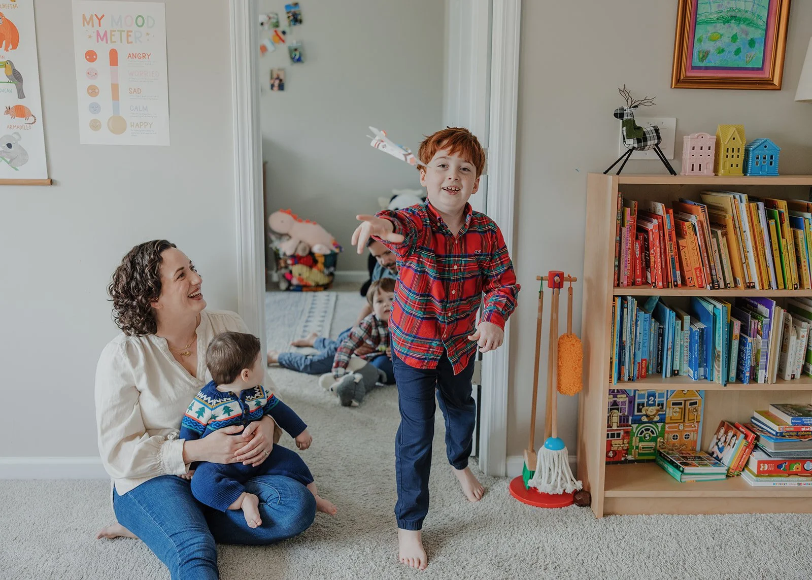 Mother sitting on the floor holding her baby while her young son runs toward the camera during a relaxed in-home family photo session with an autism-friendly family photographer in Raleigh.