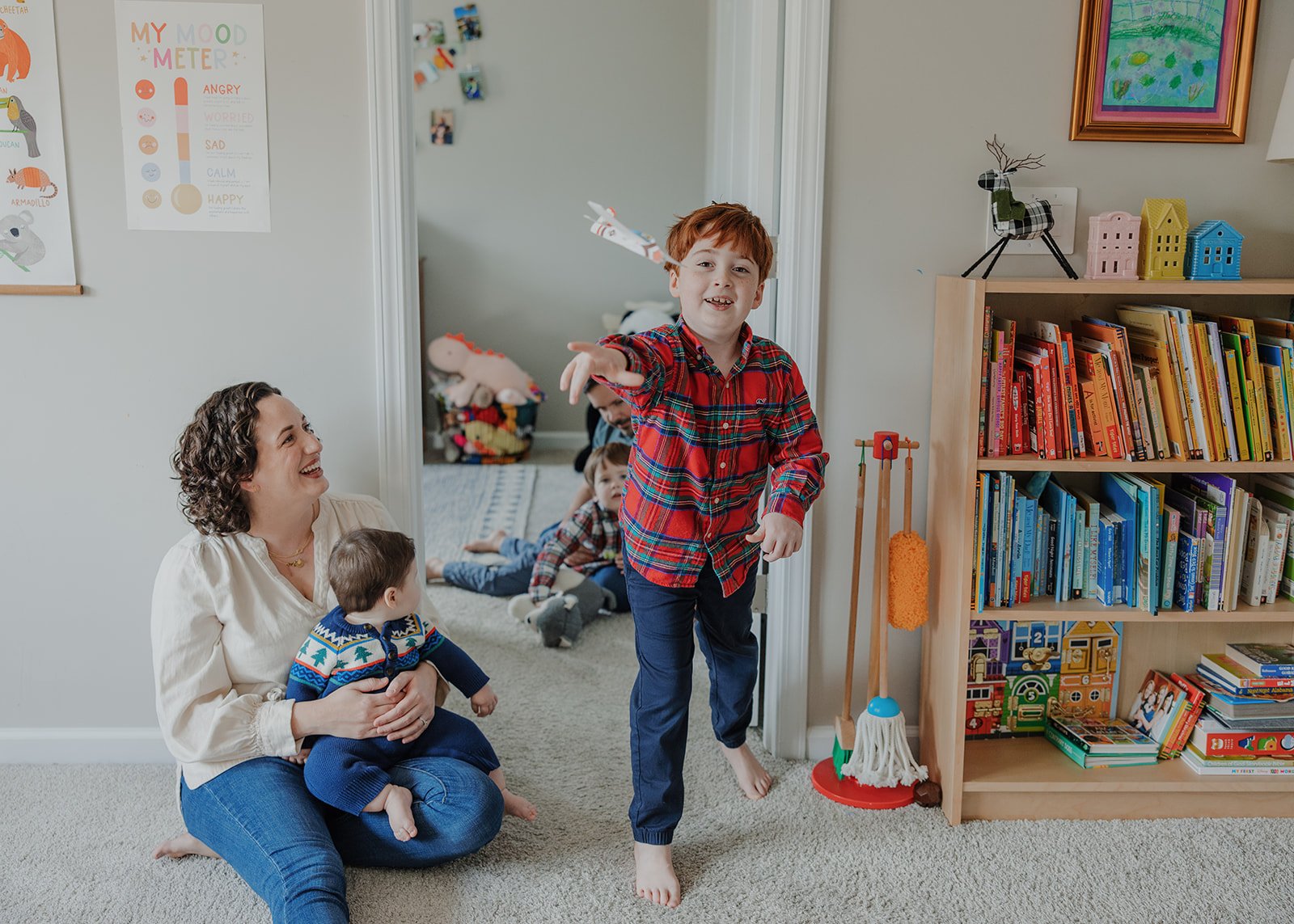 In-home family photo in Raleigh with young boy running toward the camera while mom sits on the floor holding baby and siblings play in the background, capturing a real, playful moment at home