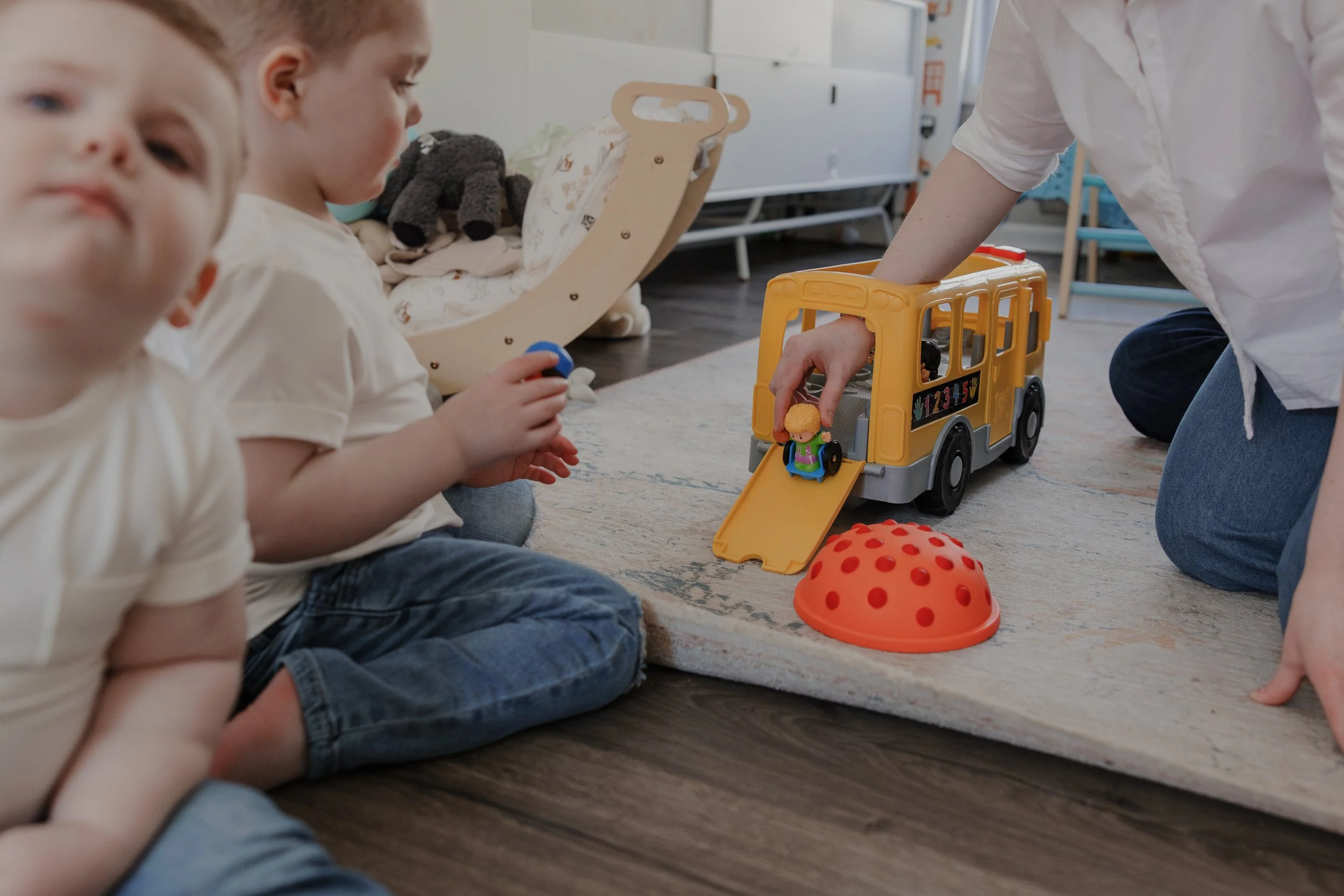 Mother plays with a toy school bus while two young brothers sit on the floor during an inclusive in-home family photography session in Durham, NC, designed for a child with ADNP Syndrome.