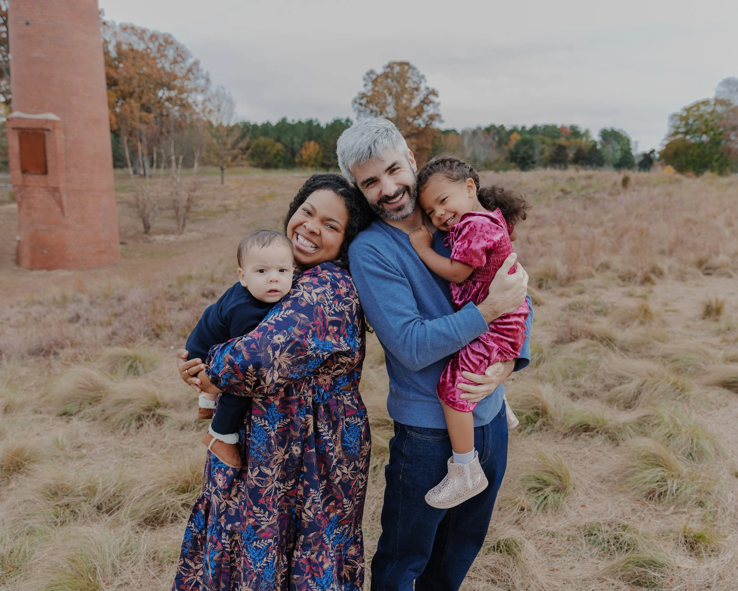 Mom and dad stand back to back while holding their kids during a fall family mini photo session at the North Carolina Museum of Art in Raleigh.