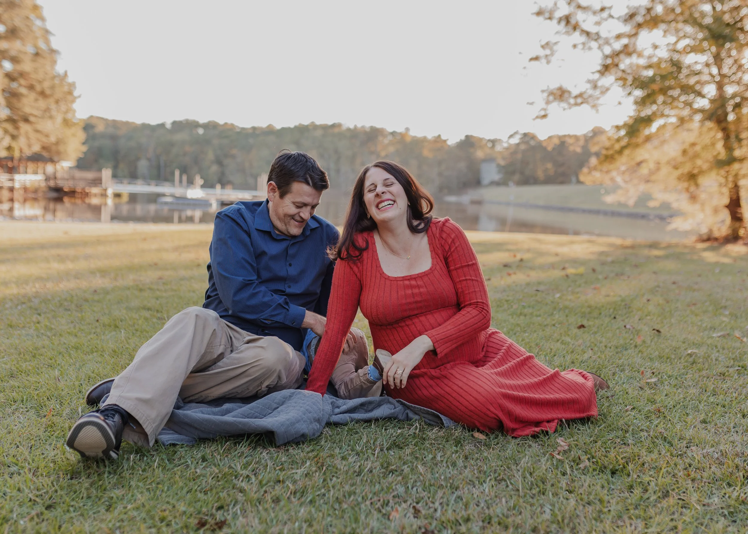 Expecting parents sitting on a blanket at Bond Park while their energetic three-year-old crawls away between them, captured during a relaxed session at Bond Park, of the sensory-friendly photo locations Raleigh and Cary families enjoy.