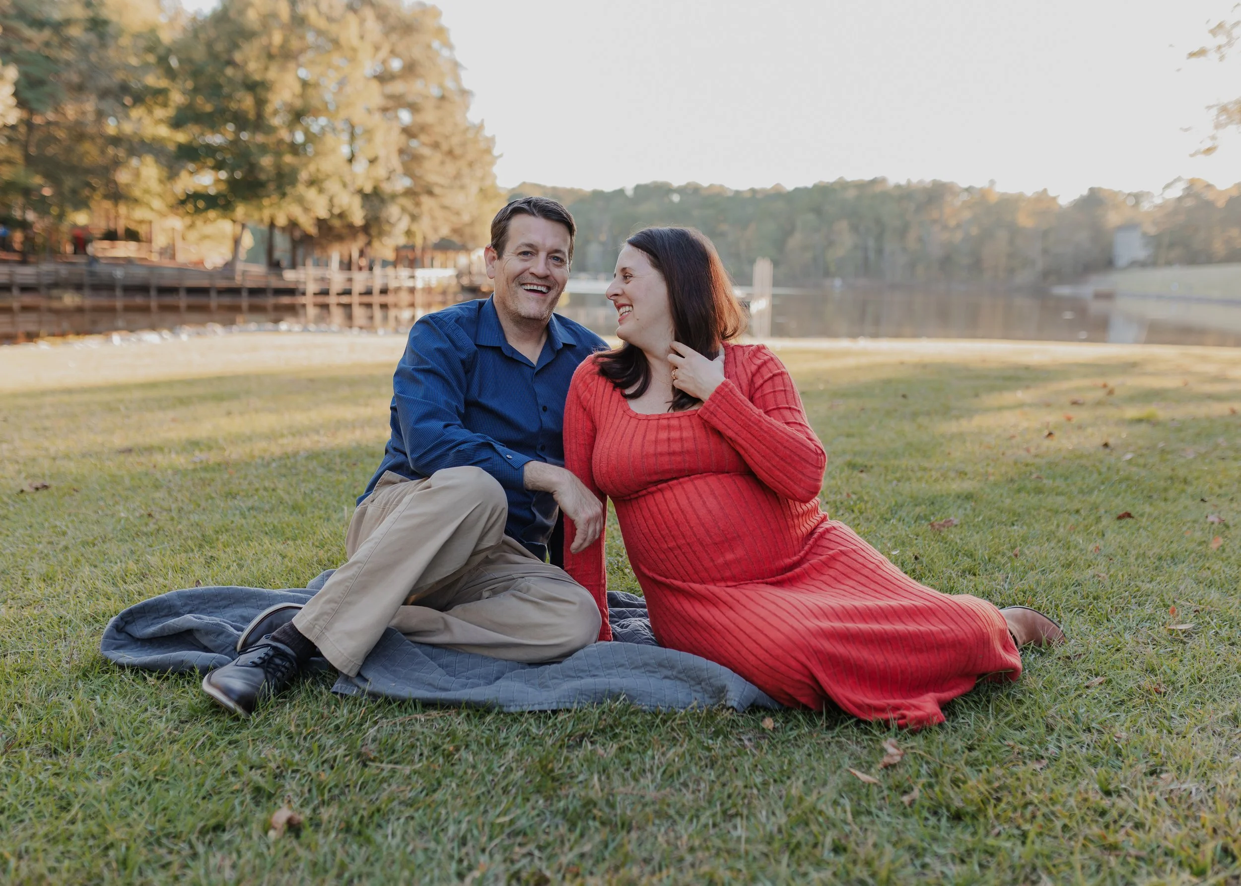 Pregnant woman sits on grass and laughs with partner at Bond Park in Cary, NC
