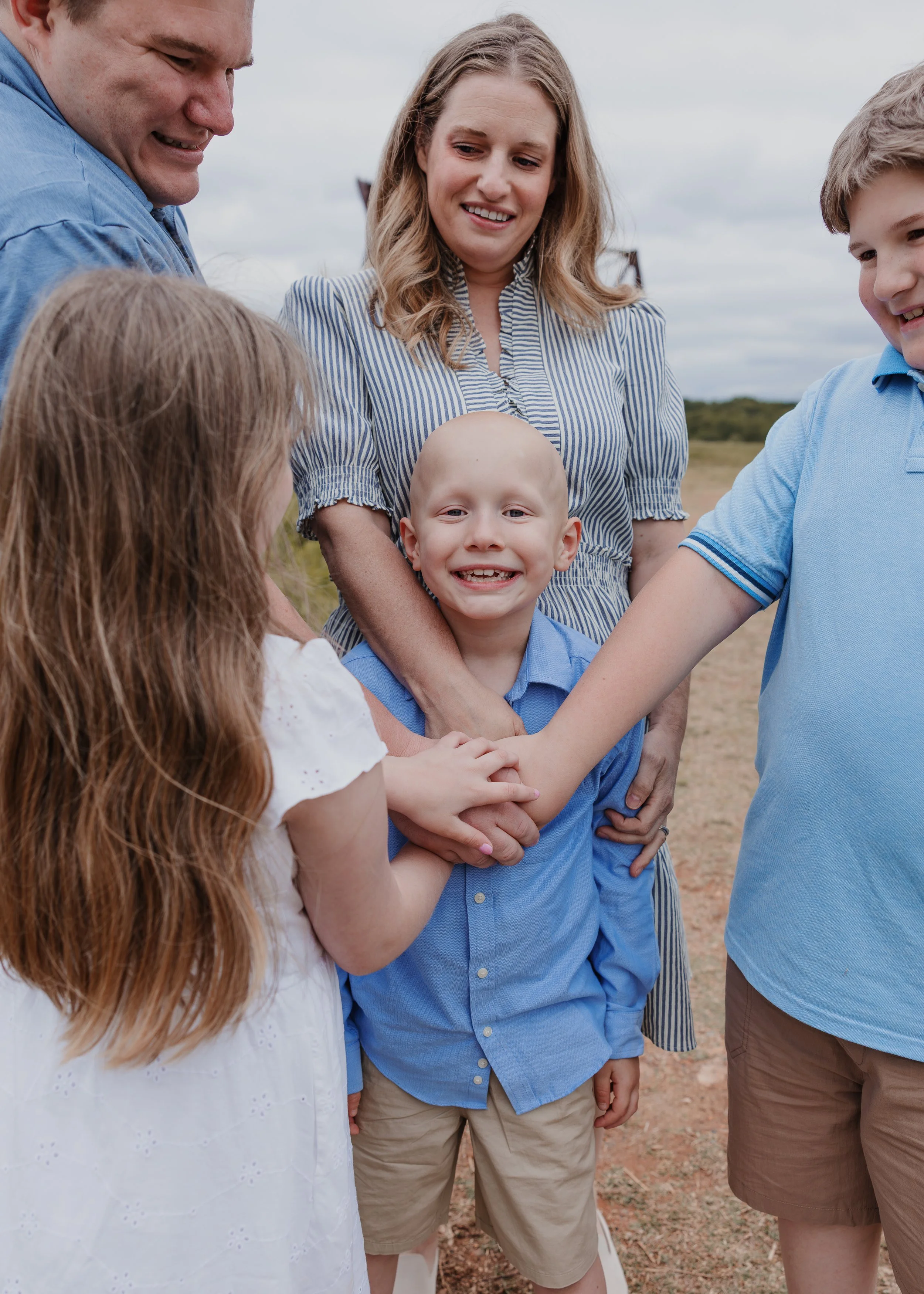 A joyful child with alopecia smiles as his family gathers around him and places their hands on his heart during a child-led family photo session at the North Carolina Museum of Art in Raleigh, NC.