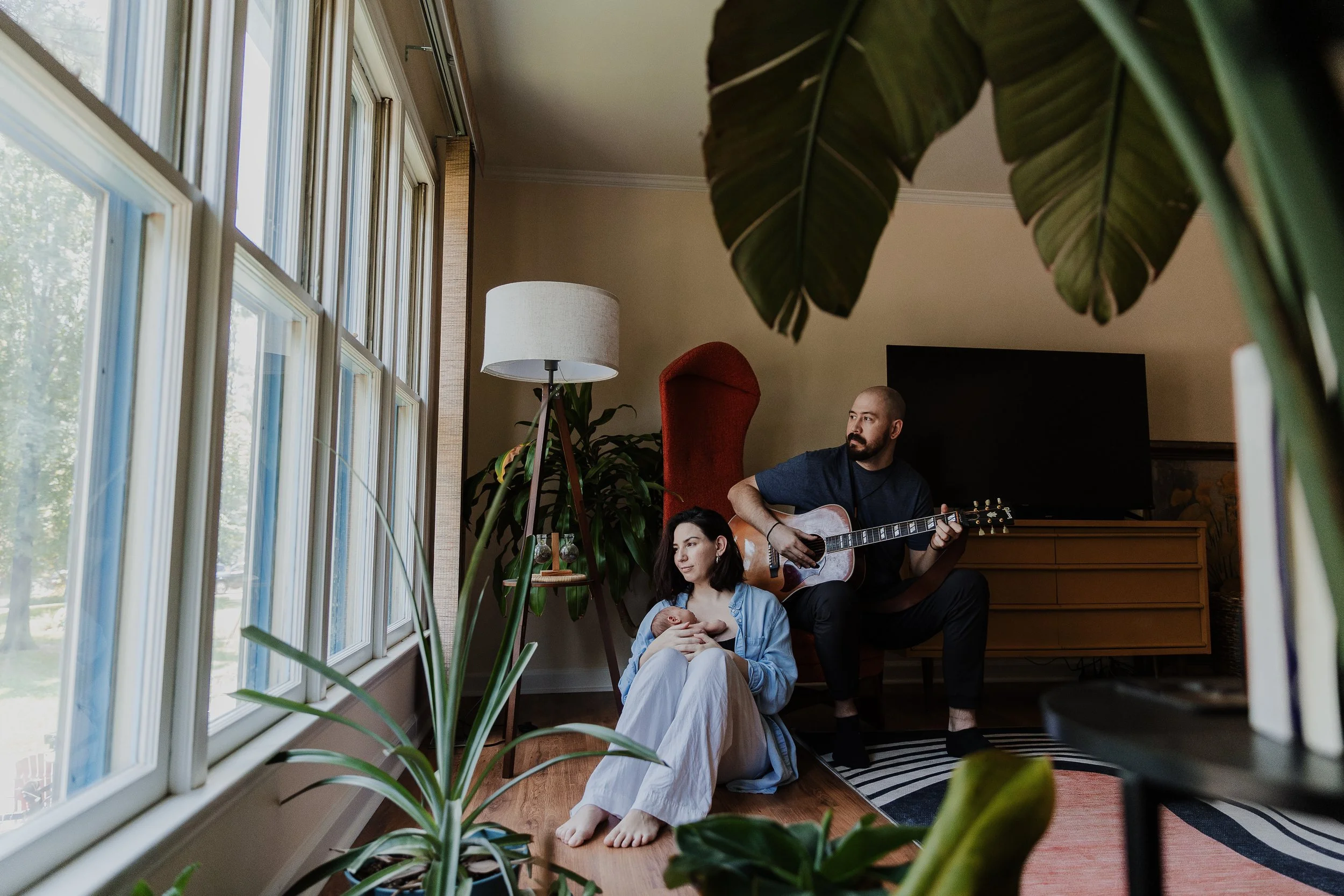 Mother sitting by window holding newborn baby while husband plays guitar during a relaxed in-home newborn photography session in Raleigh.