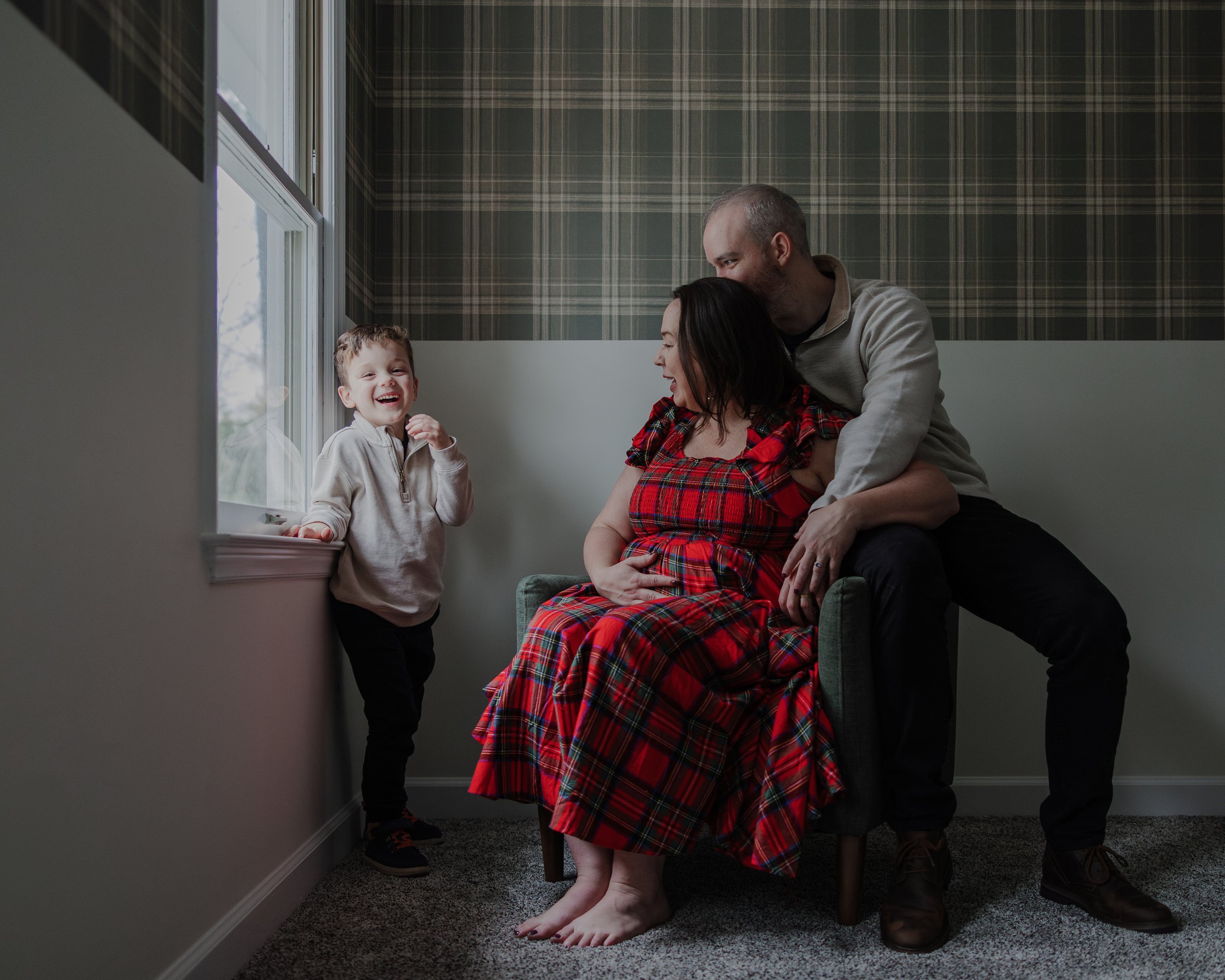 Professional maternity photo session in Durham with a pregnant mother in a red dress sitting by a window, her partner leaning in close, and their young child laughing beside them in a cozy, natural-light home.