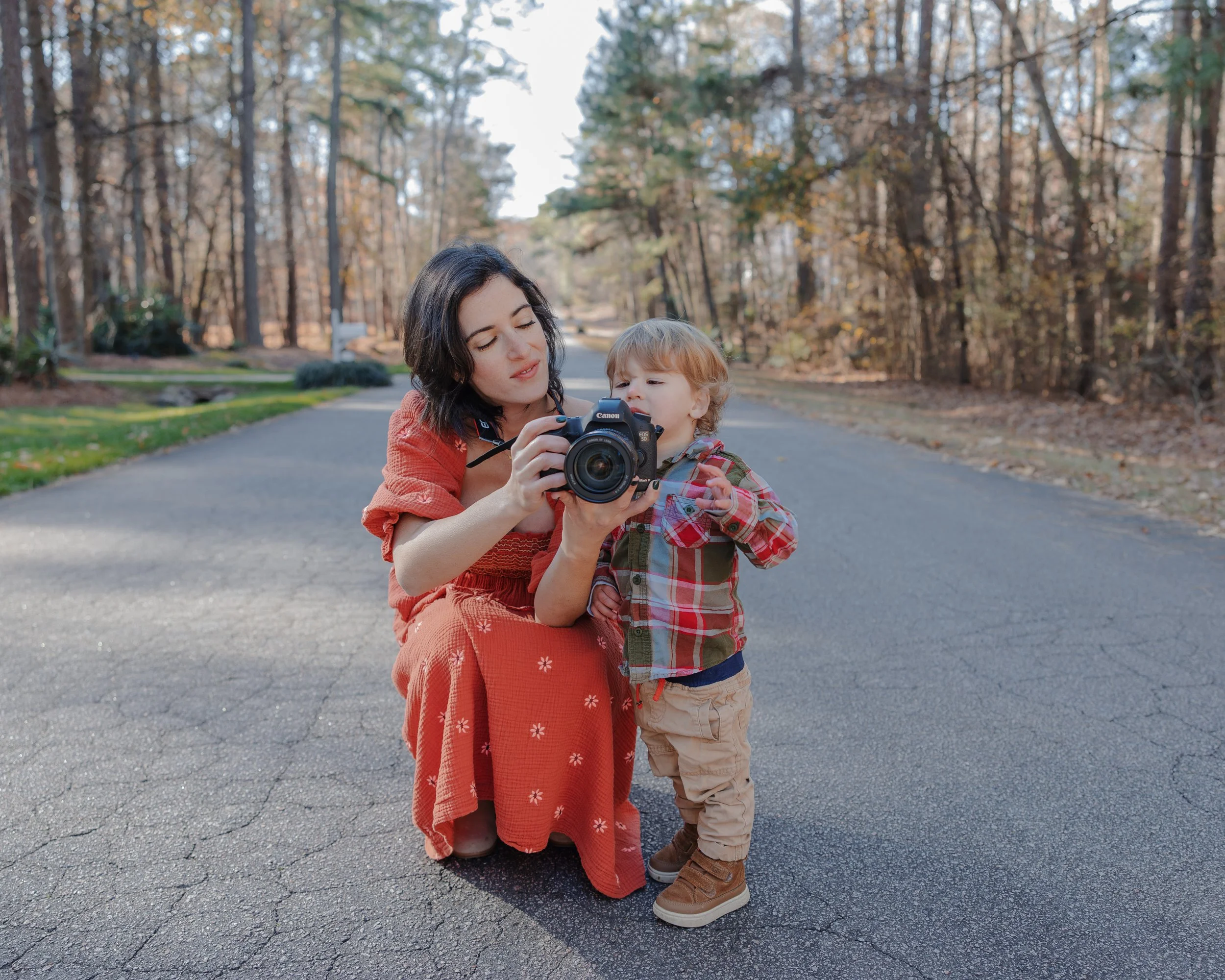 A Raleigh school photographer in an orange dress kneeling on a paved path to show a young boy in a plaid shirt his portrait on the back of a professional camera.