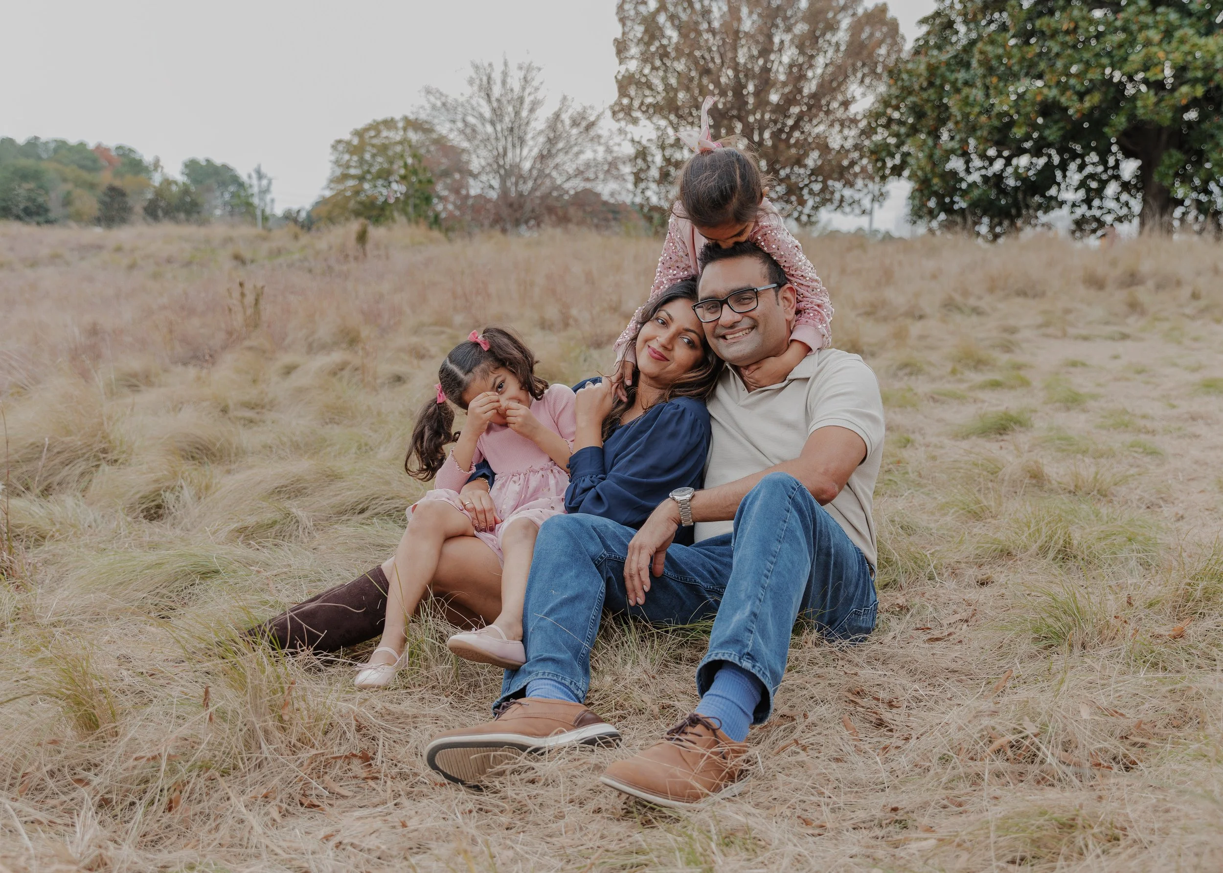 Parents sitting in a field at the NC Museum of Art Park with their daughters climbing and snuggling around them during a playful family moment.