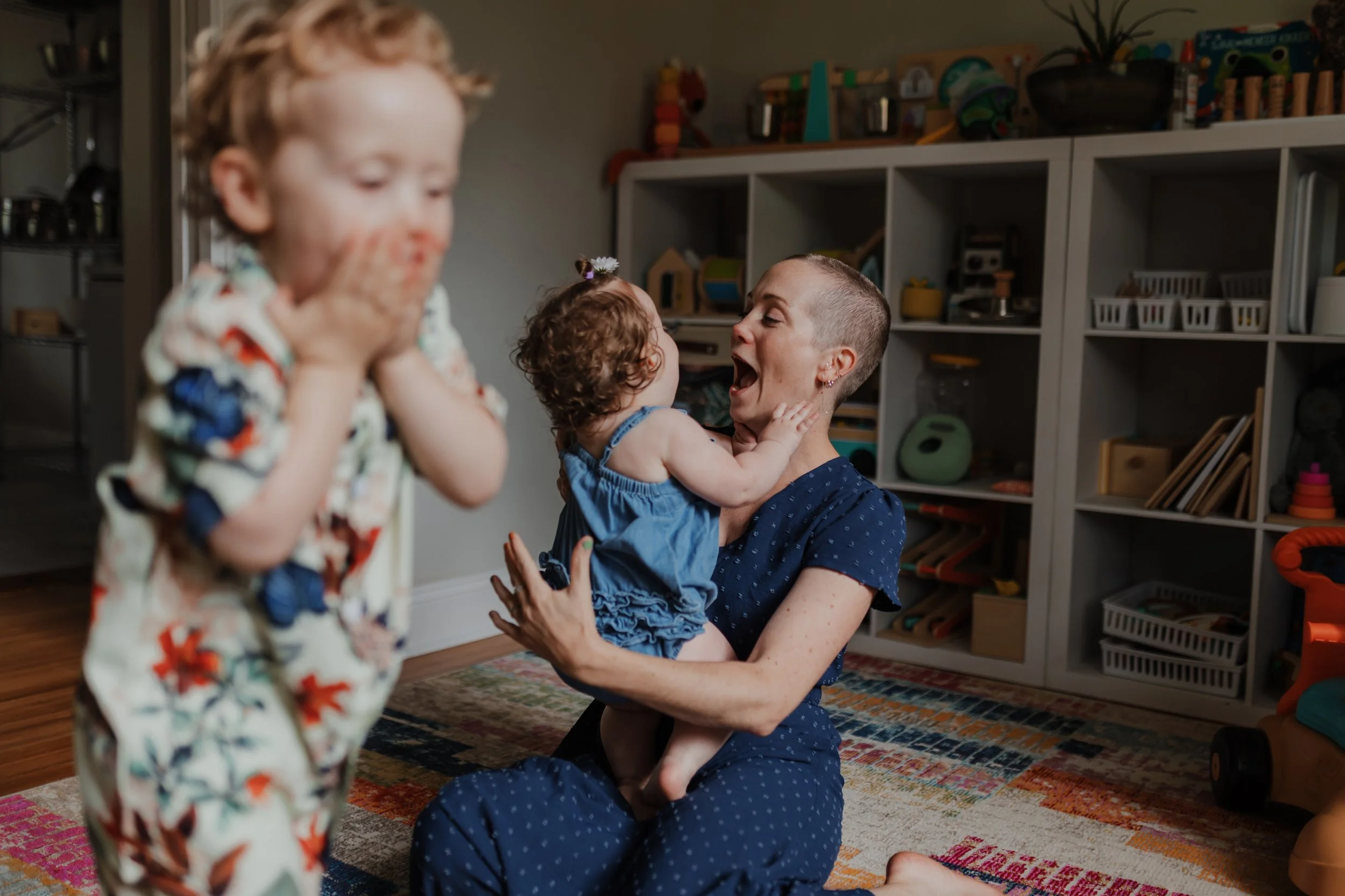 Mom kisses baby while 5-year-old boy runs around during a cozy in-home family photo session in Wake Forest, NC.