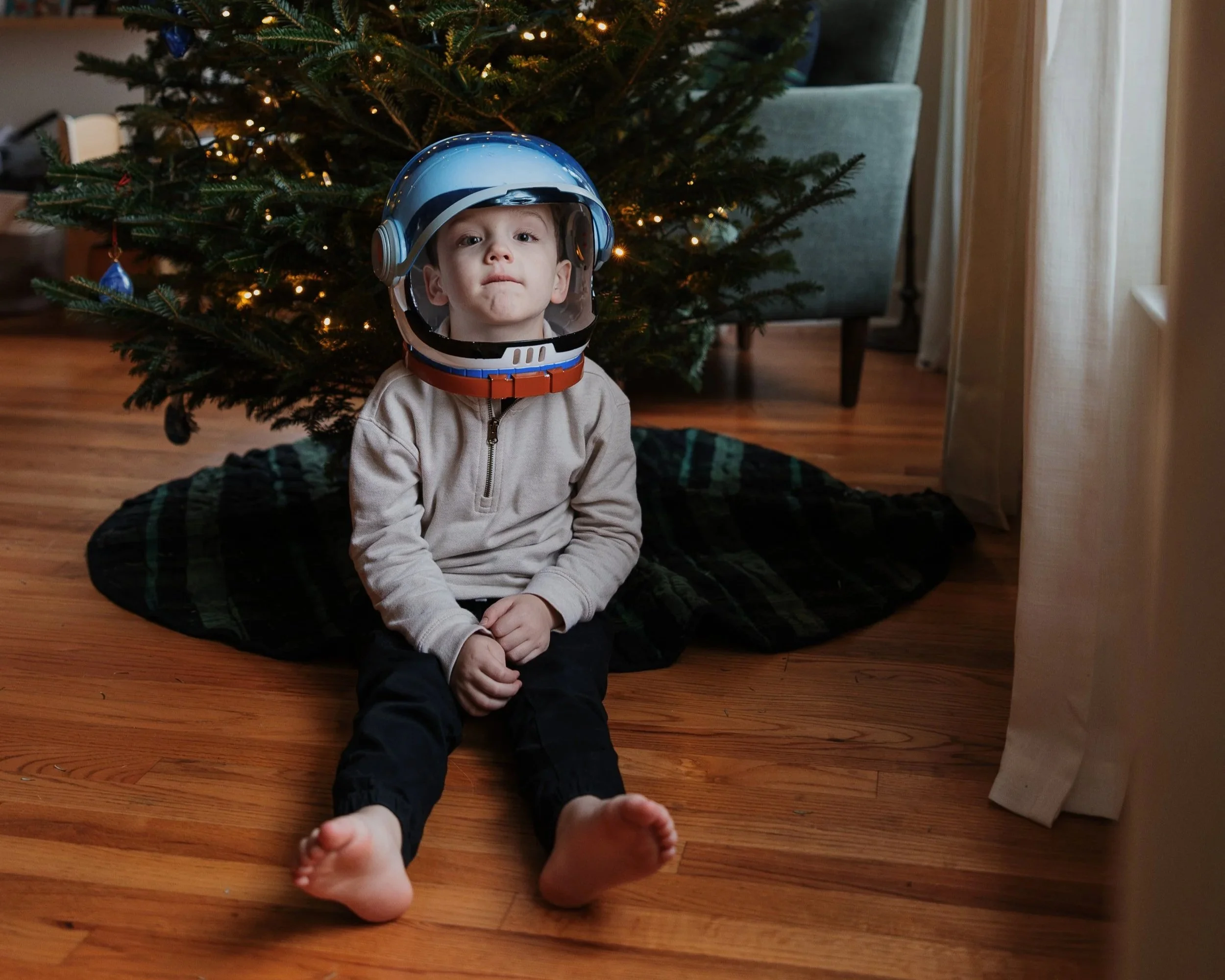 Young boy wearing an astronaut helmet sits in front of his family's Christmas tree during a full-length family photography session in Durham, NC.