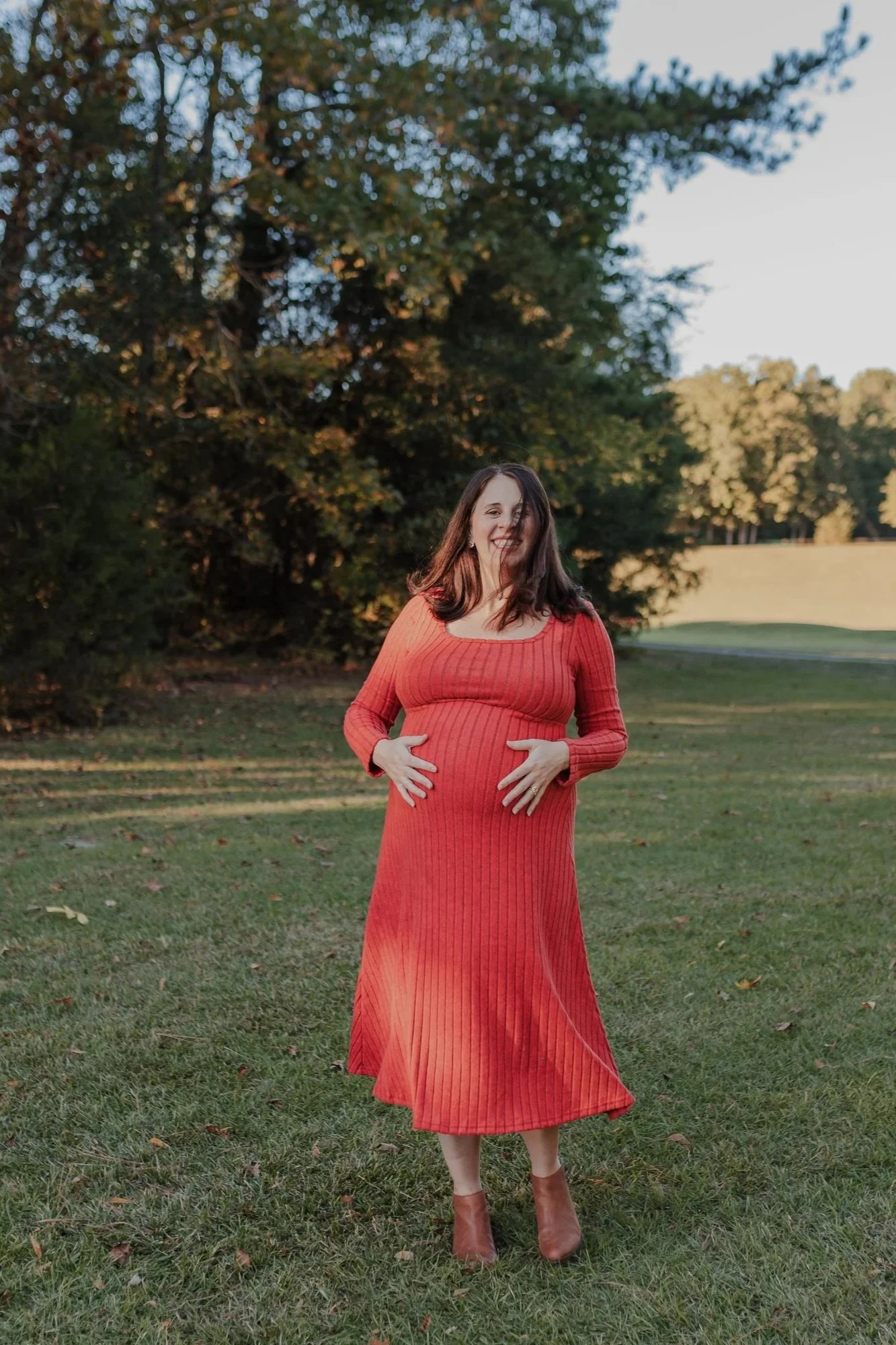 Pregnant woman in a red dress standing in an open field at Joyner Park in Raleigh, NC during a natural light maternity photo session