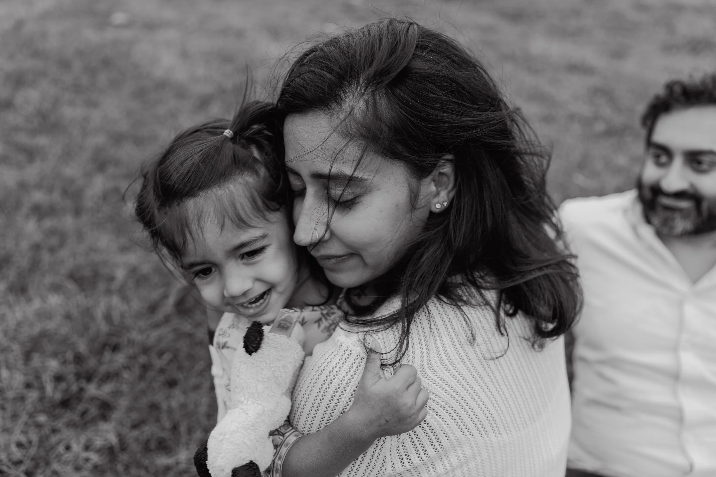 Mom hugs three-year-old daughter while dad watches during a professional family photo session at NCMA in Raleigh, NC.