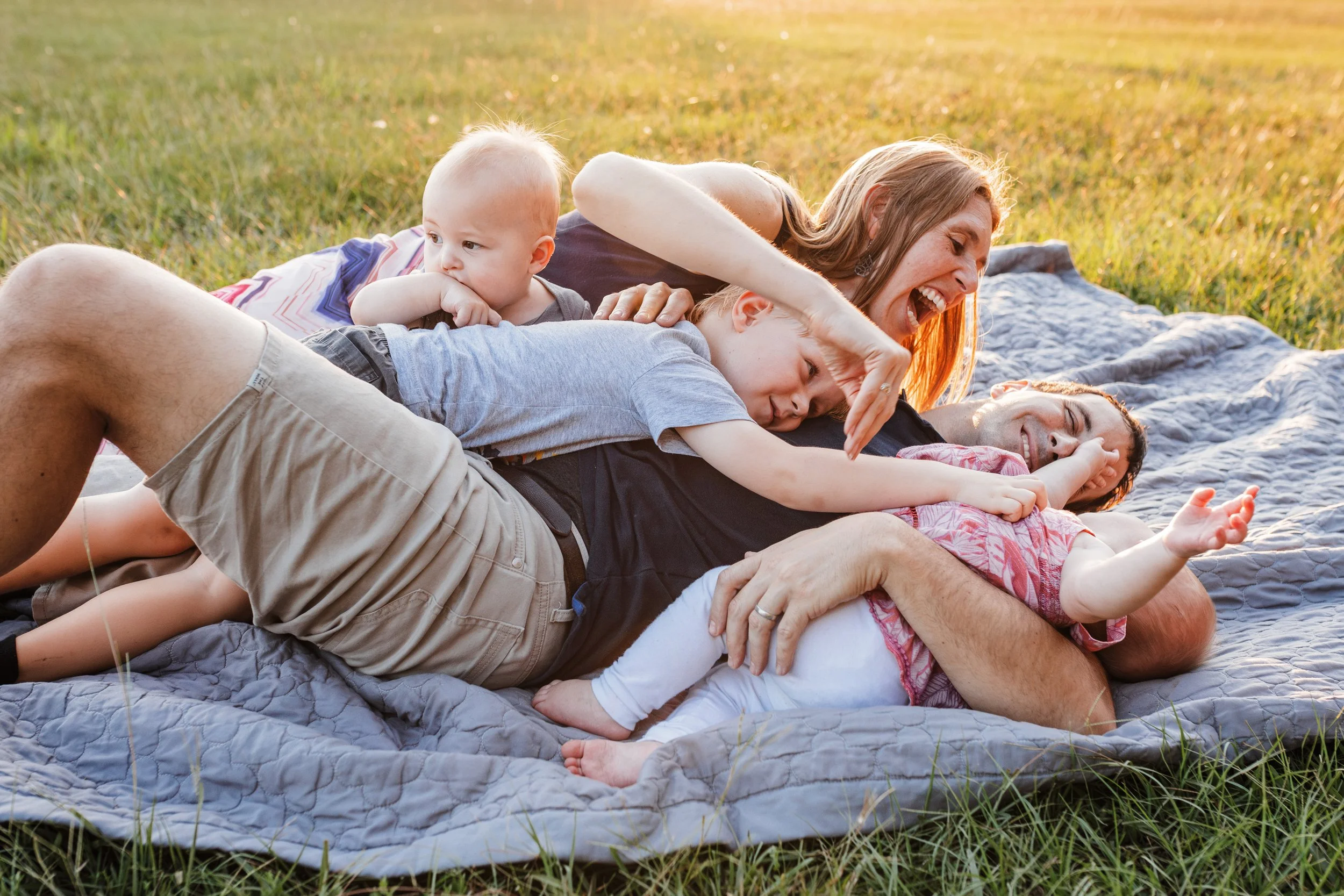 Parents and young children cuddling and laughing on a blanket in the grass during golden hour at Lake Crabtree County Park in Cary.