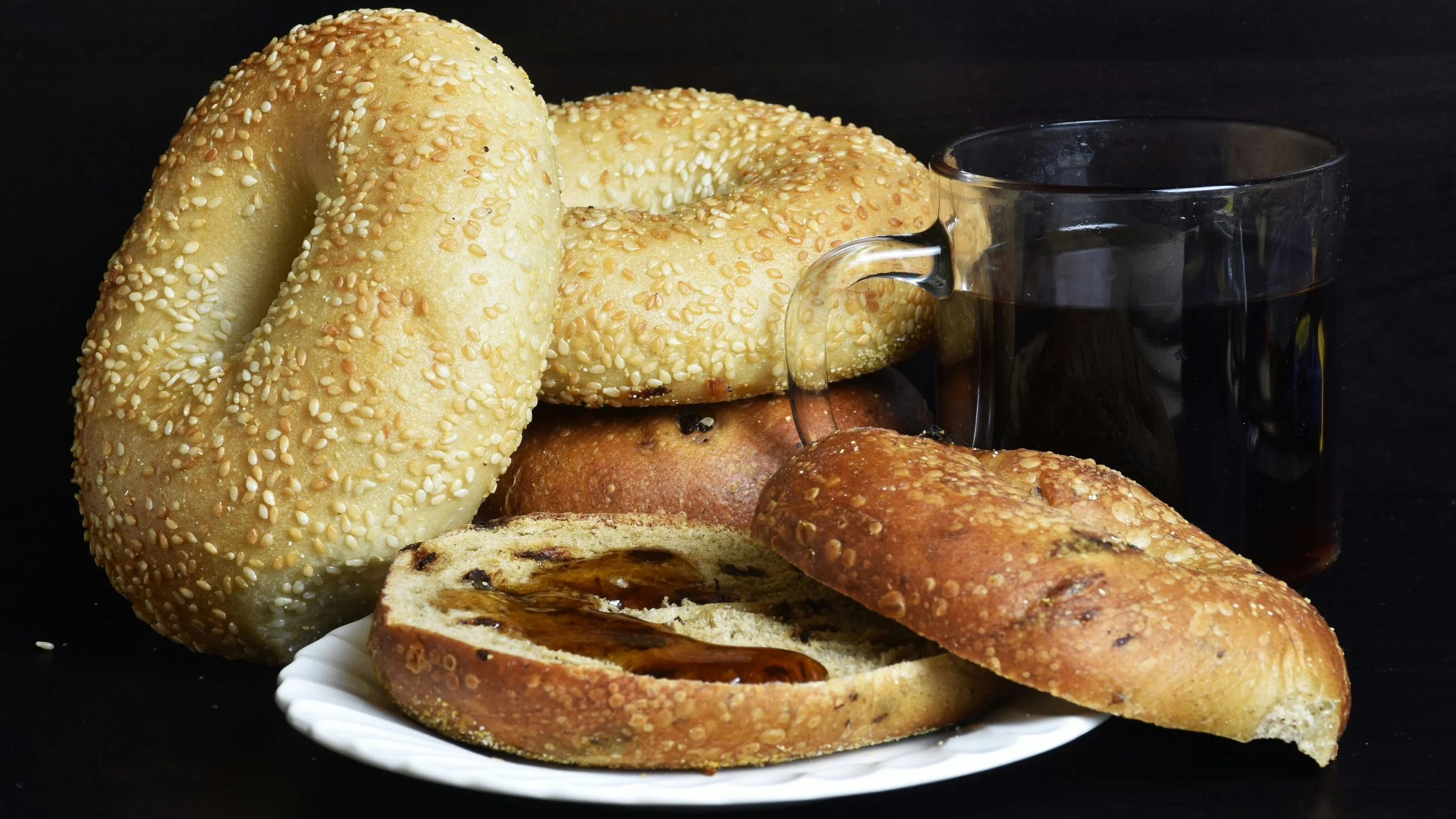 Several different bagels, one on a plate with some kind of smear on it and a clear cup of black coffee