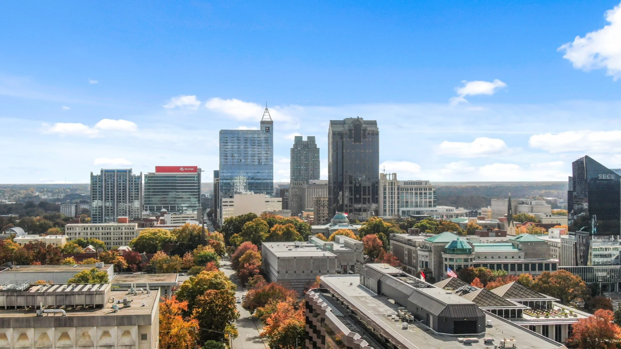 Skyline view of a city with tall skyscrapers, trees with autumn-colored leaves, and a partly cloudy blue sky.