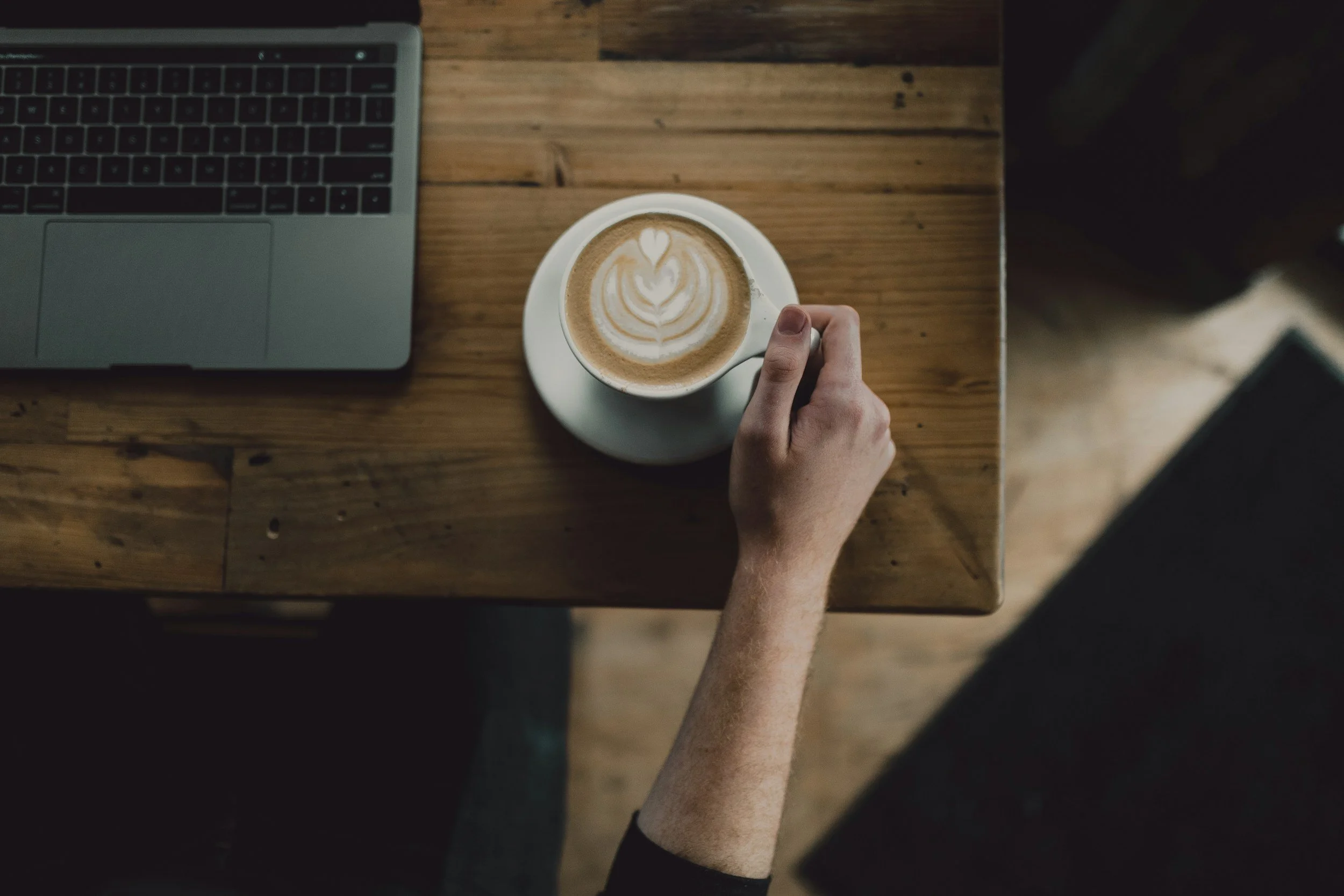 A person's hand holding a latte with latte art on top, placed on a white saucer on a wooden table next to a laptop.