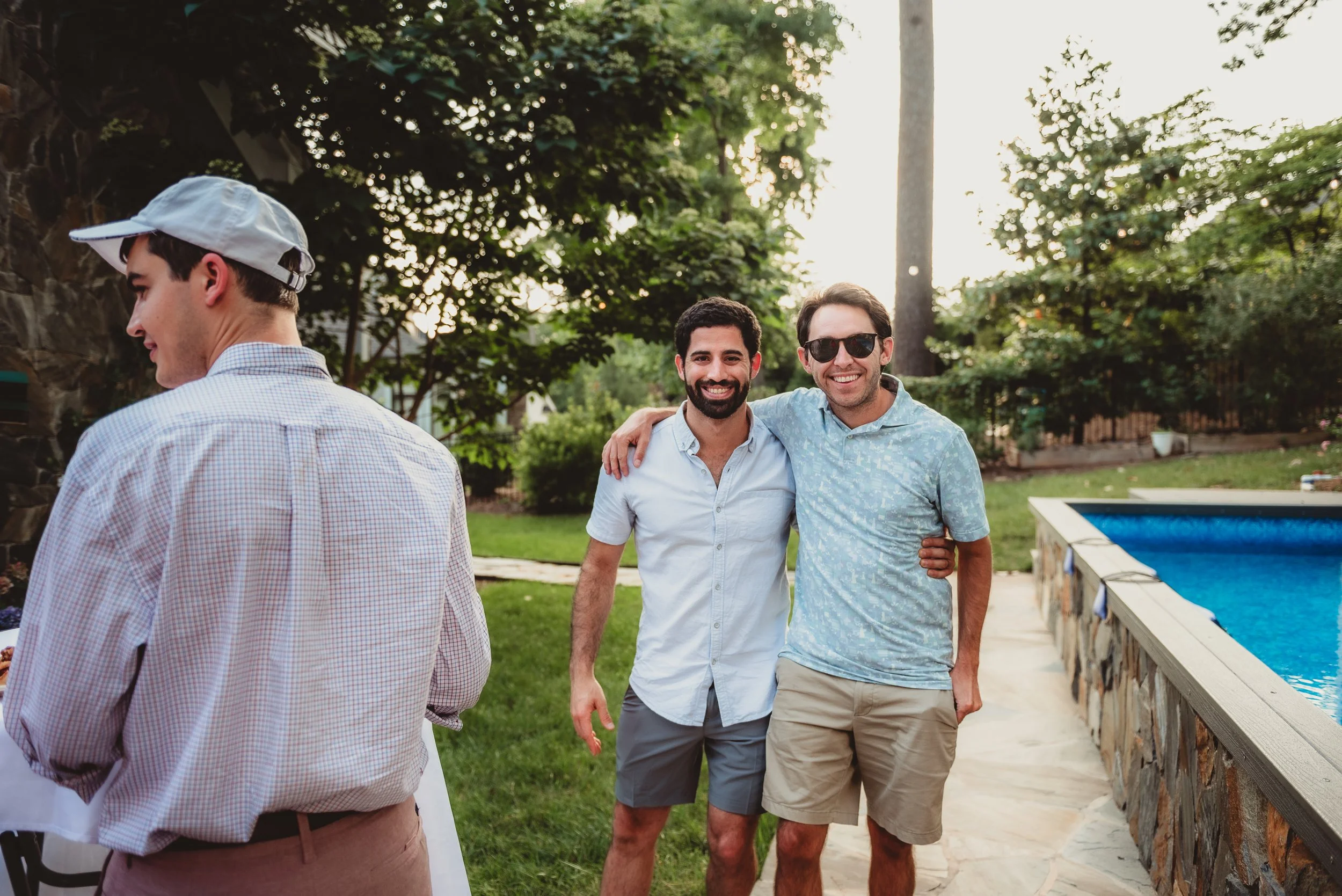 Two men smiling and arm-in-arm outdoors near a swimming pool during a gathering, with a third person partly visible on the left side of the image.