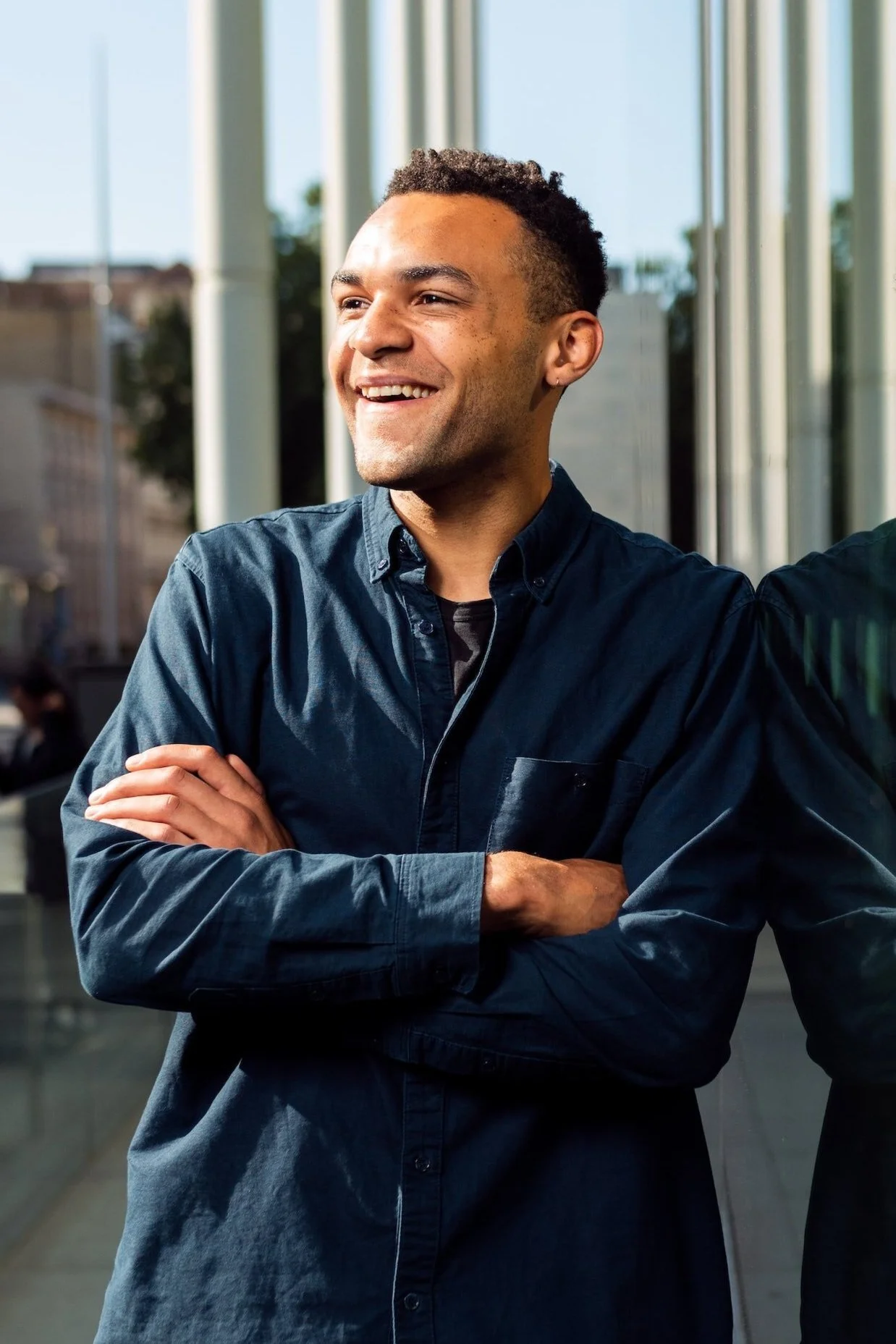A smiling person in a dark blue shirt crossing their arms, standing outdoors next to a reflective glass surface.