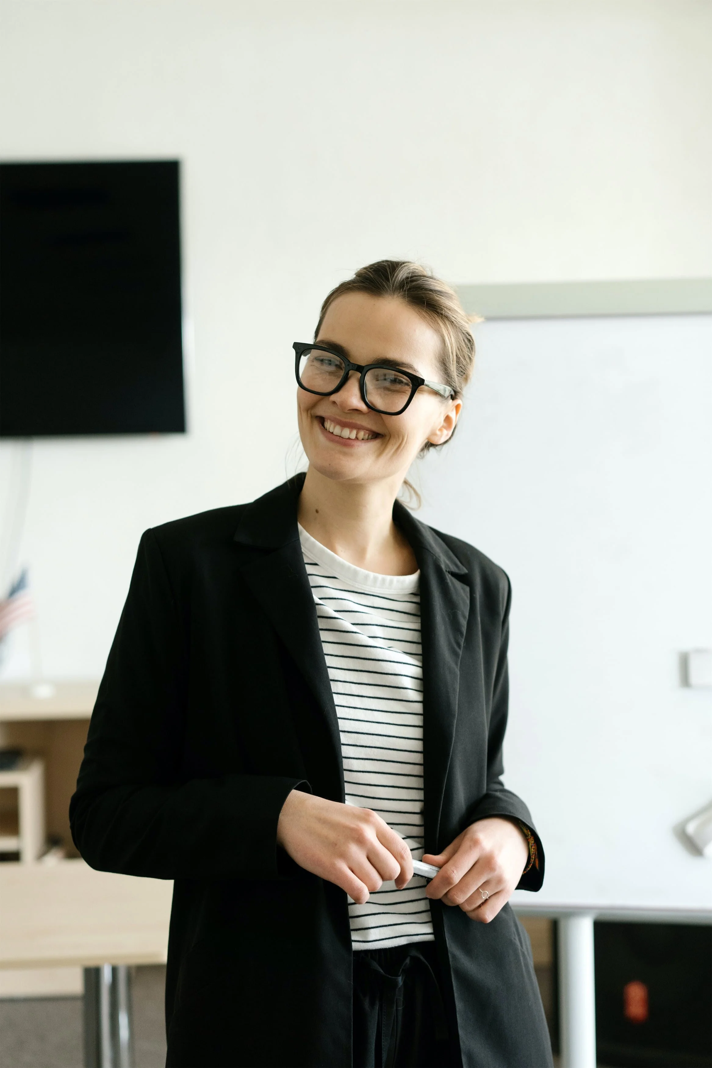 Person smiling in a black blazer and striped shirt, standing in a classroom with a whiteboard and an American flag in the background.