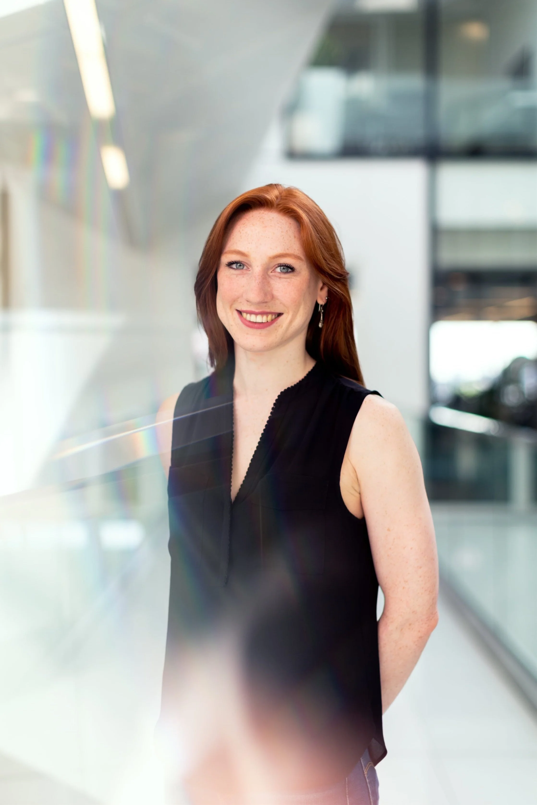 A woman with red hair wearing a black sleeveless top smiles in a bright, modern interior setting with glass walls and soft light reflections.