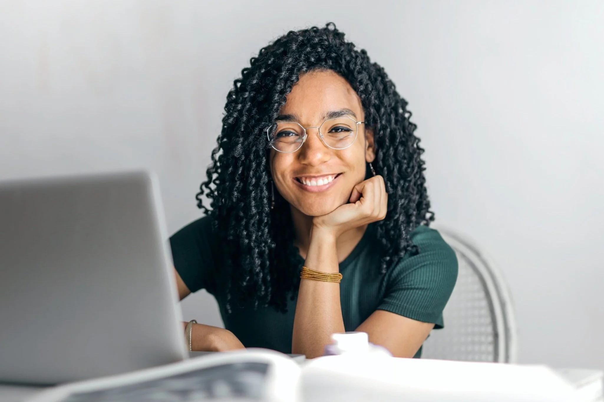 Person with curly hair smiling, sitting at a table with a laptop and books.