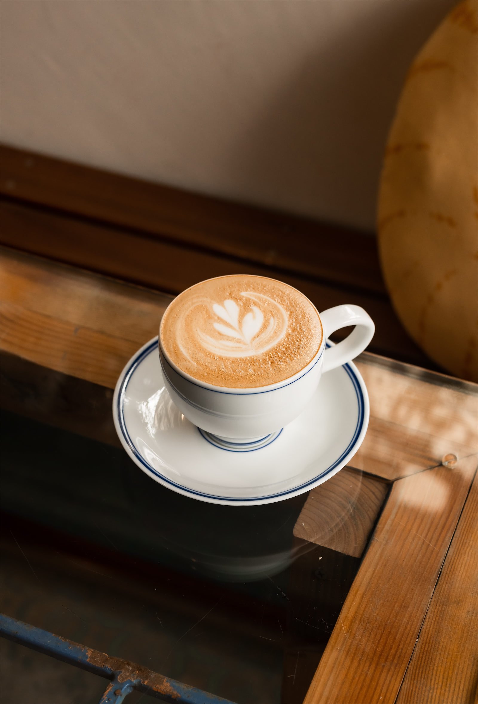 A cup of latte with latte art in a white cup with a blue stripe on a matching saucer, placed on a wooden table.