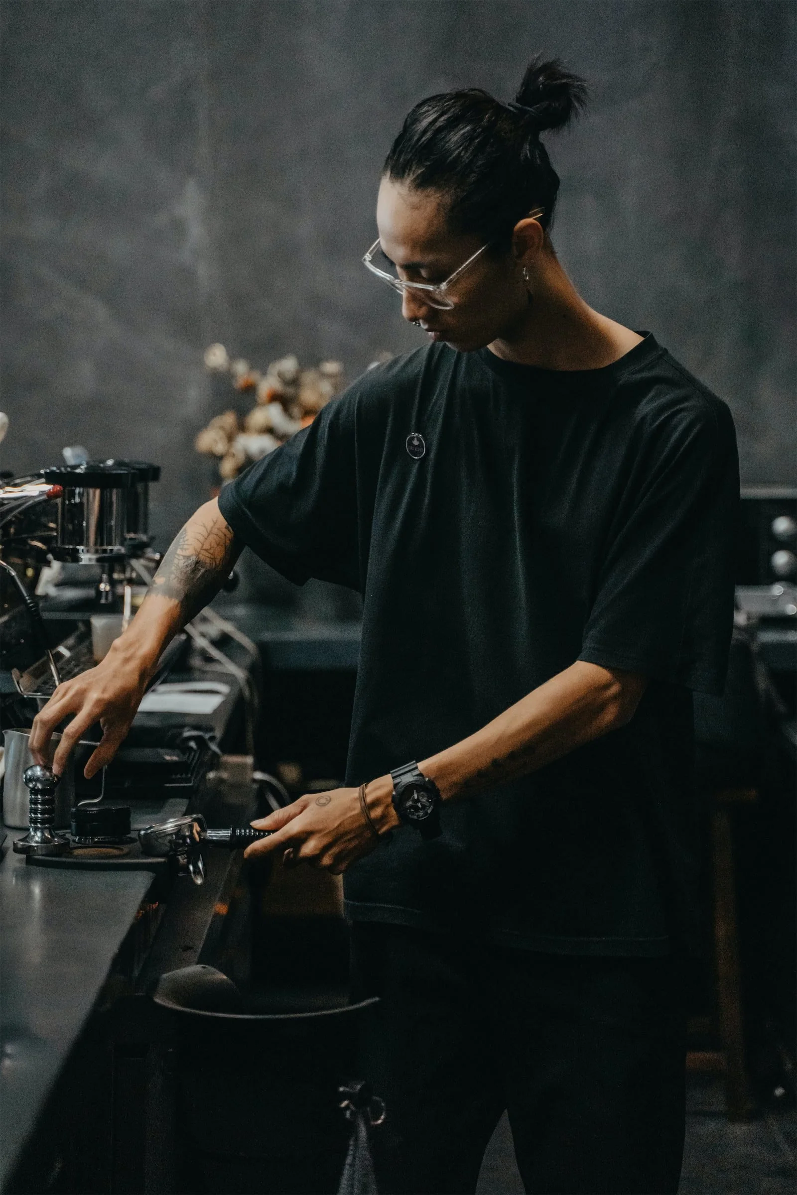 A person with tattoos wearing glasses, a black T-shirt, and a wristwatch, working on brewing coffee in a coffee shop or cafe with a dark, industrial interior.