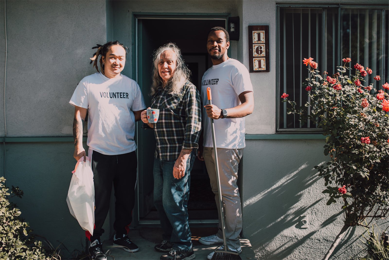 Three volunteers and an elderly woman standing at the doorway of a house, smiling, with some gardening tools and a mug in hand.