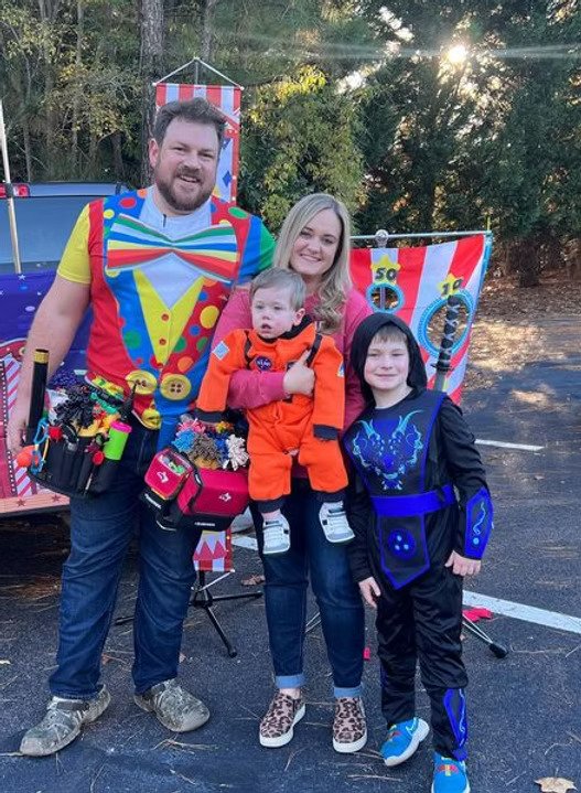 A family dressed in colorful costumes stands outdoors in front of a carnival game booth. The man is dressed as a clown with a multicolored bow tie and suspenders, the woman is holding a child dressed as an astronaut, and another child is dressed as a superhero in a dark costume with blue accents. They are smiling for the camera.