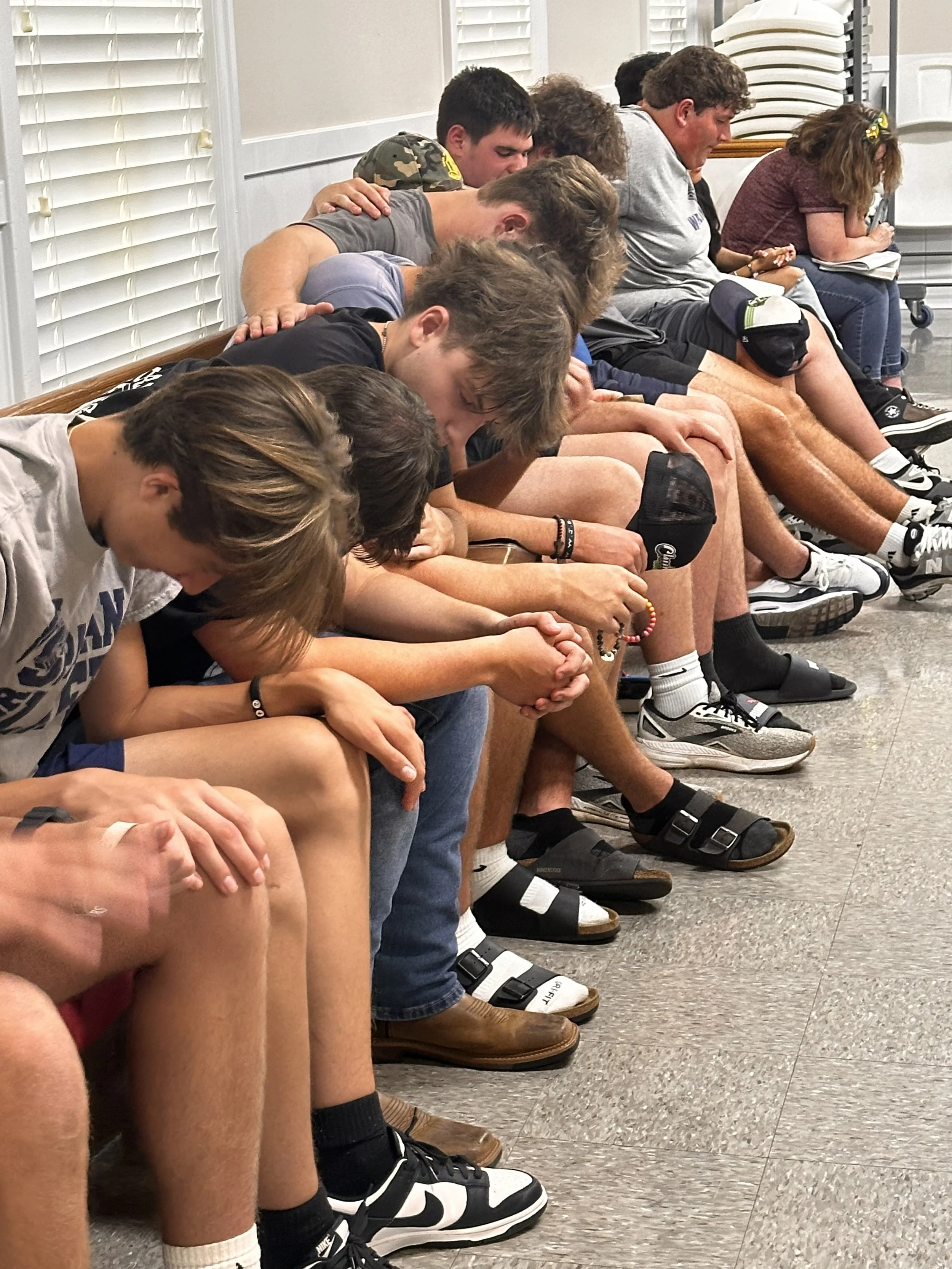 A group of young people sitting on a bench with their heads bowed and eyes closed, appearing to be in prayer or reflection, inside a room with white blinds and stacked chairs in the background.