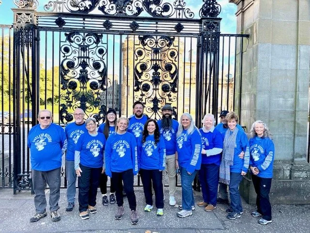 Group of eleven people standing in front of a large, ornate black iron gate with a stone wall to the right. They are all wearing matching blue shirts with white text and graphics, outdoors on a sunny day.