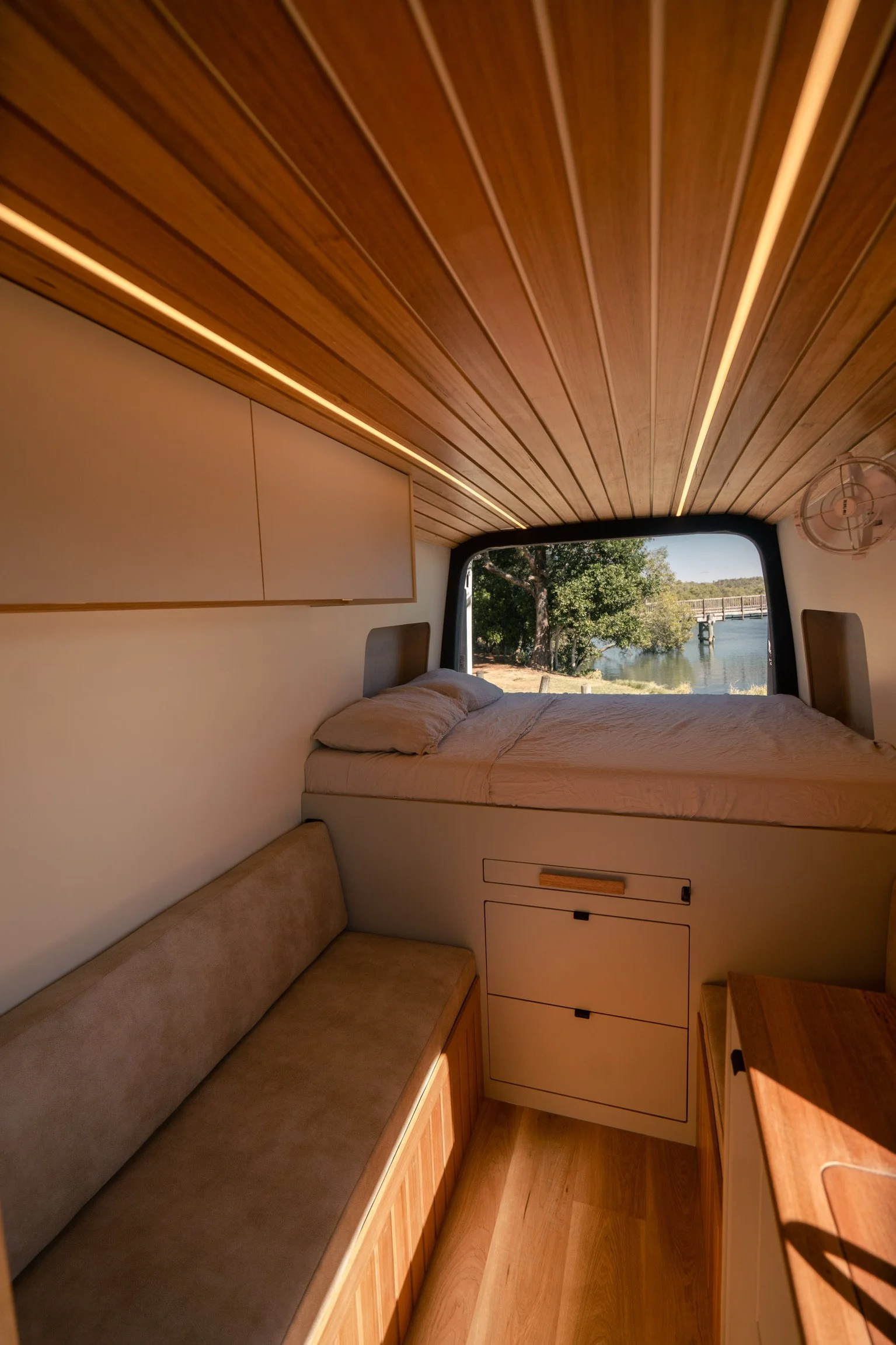 Interior of a small, modern camper with a wooden ceiling, a bed near a large window showing an outdoor river scene, and built-in storage.