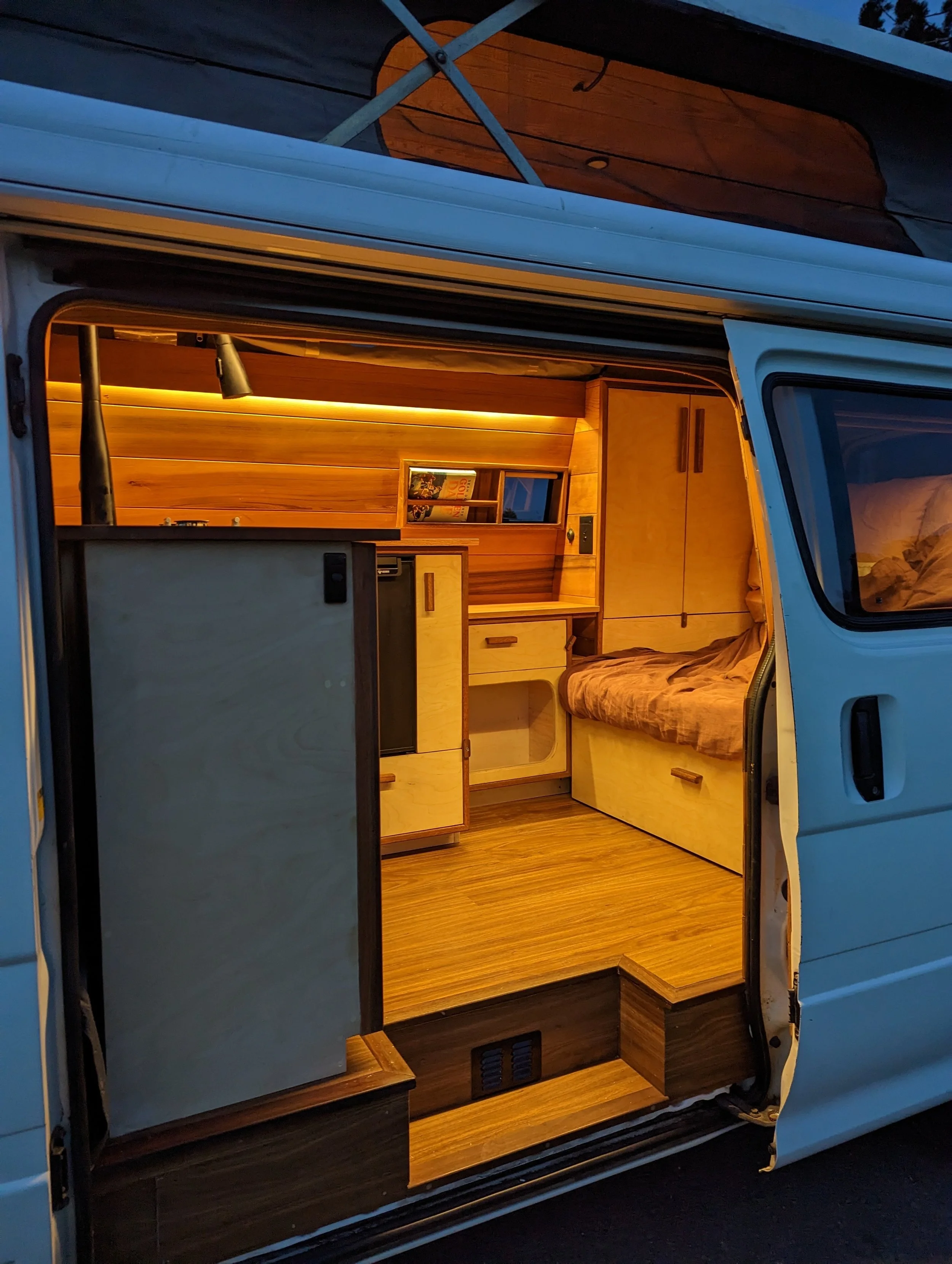 Interior of a converted camper van showcasing wooden cabinets, a small refrigerator, a bed with a blanket, and warm lighting.