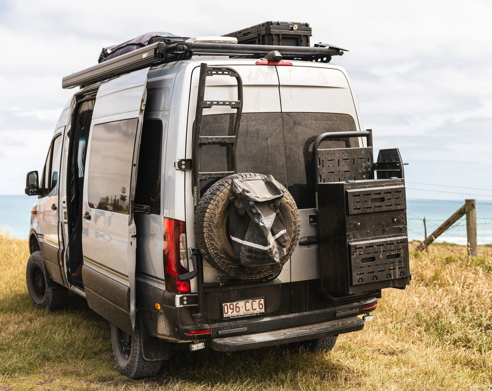 A gray camper van parked on grass near the coast with outdoor gear on its roof and rear, including a spare tire, storage boxes, and a ladder.
