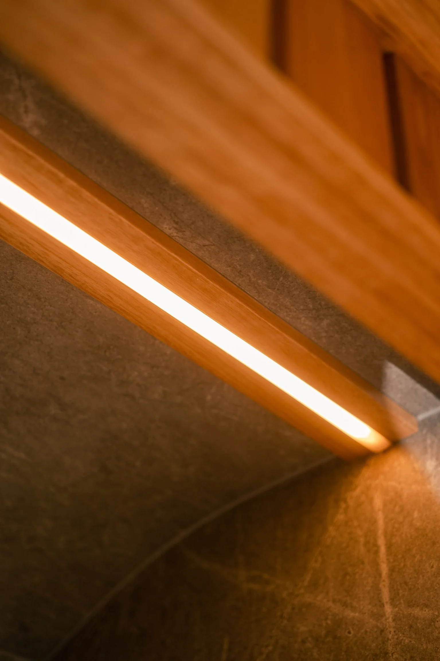 Close-up of a wooden LED strip light under a wooden cabinet, illuminating the dark brown tiled floor below.