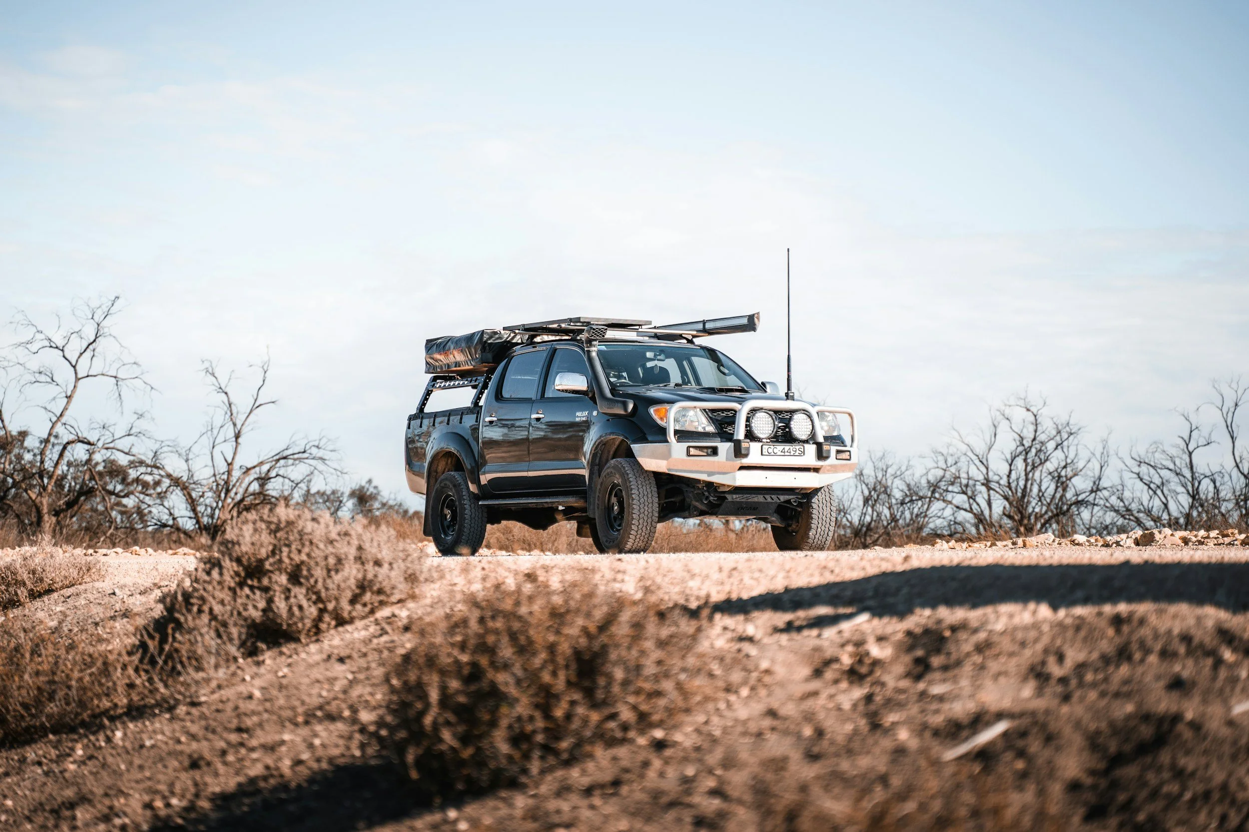 A black pickup truck is parked on a dirt trail in a barren landscape with leafless trees and a partly cloudy sky.