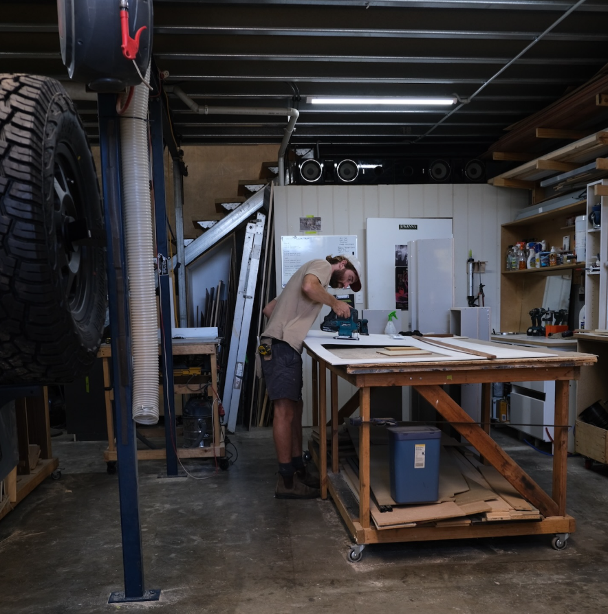 A man working in a woodworking shop, using a power drill at a large workbench surrounded by tools, wood, and storage shelves.