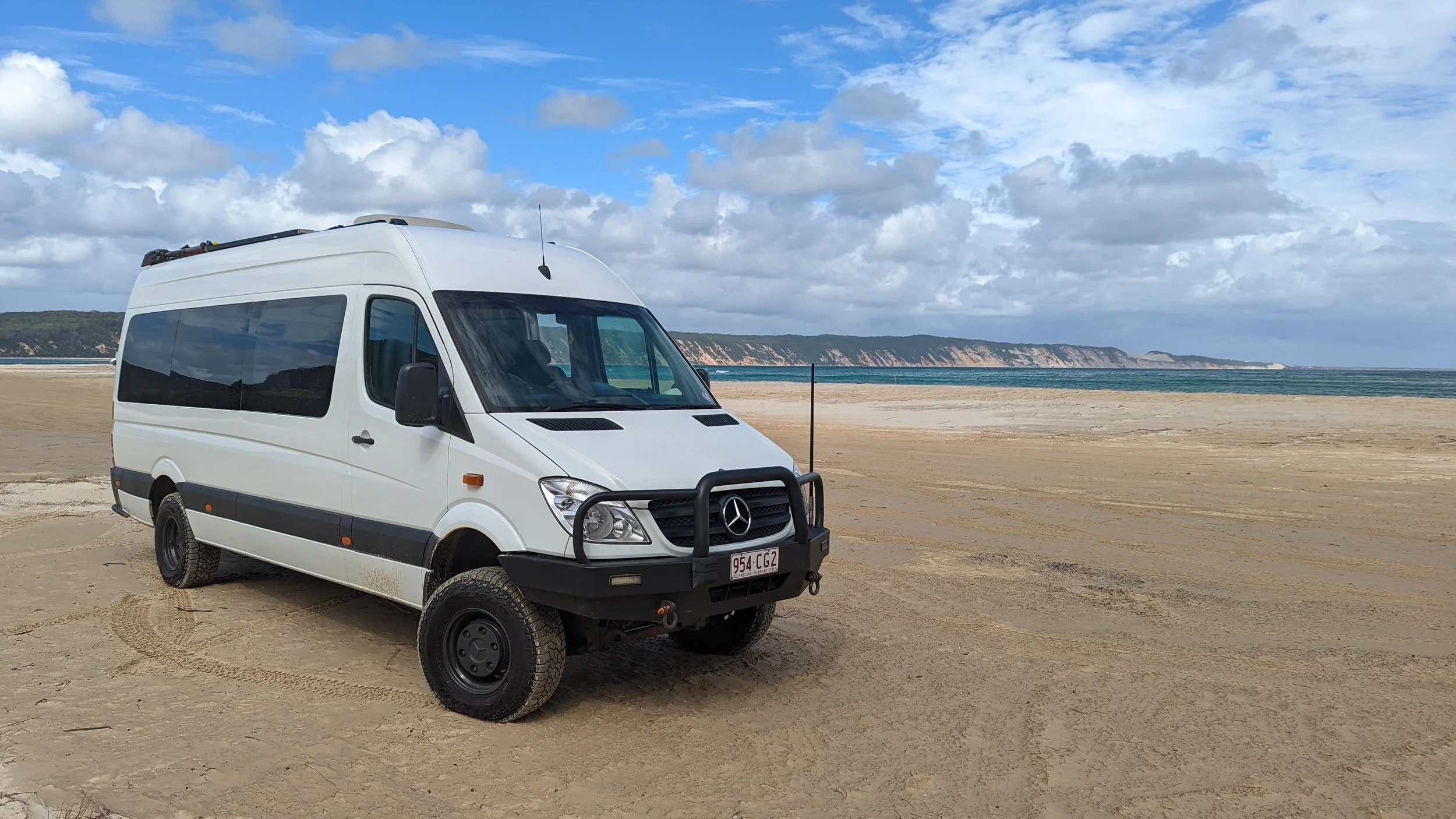 White Mercedes-Benz van parked on a sandy beach with cliffs and ocean in the background.