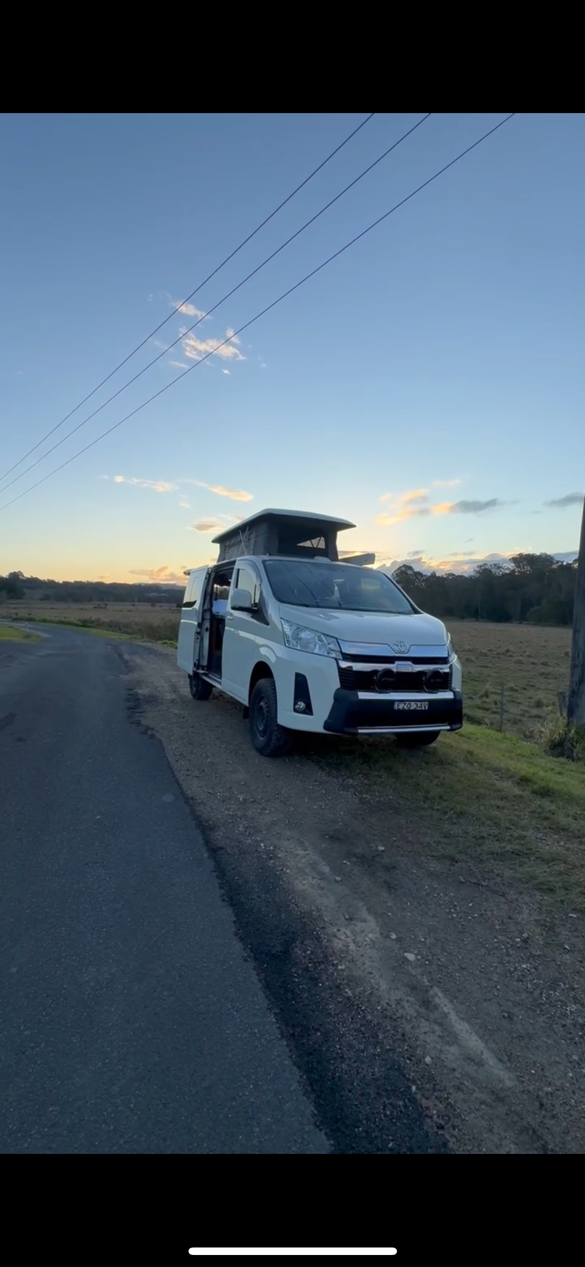 A white camper van with a rooftop tent parked beside a rural road during sunset, with open side doors and utility poles in the background.