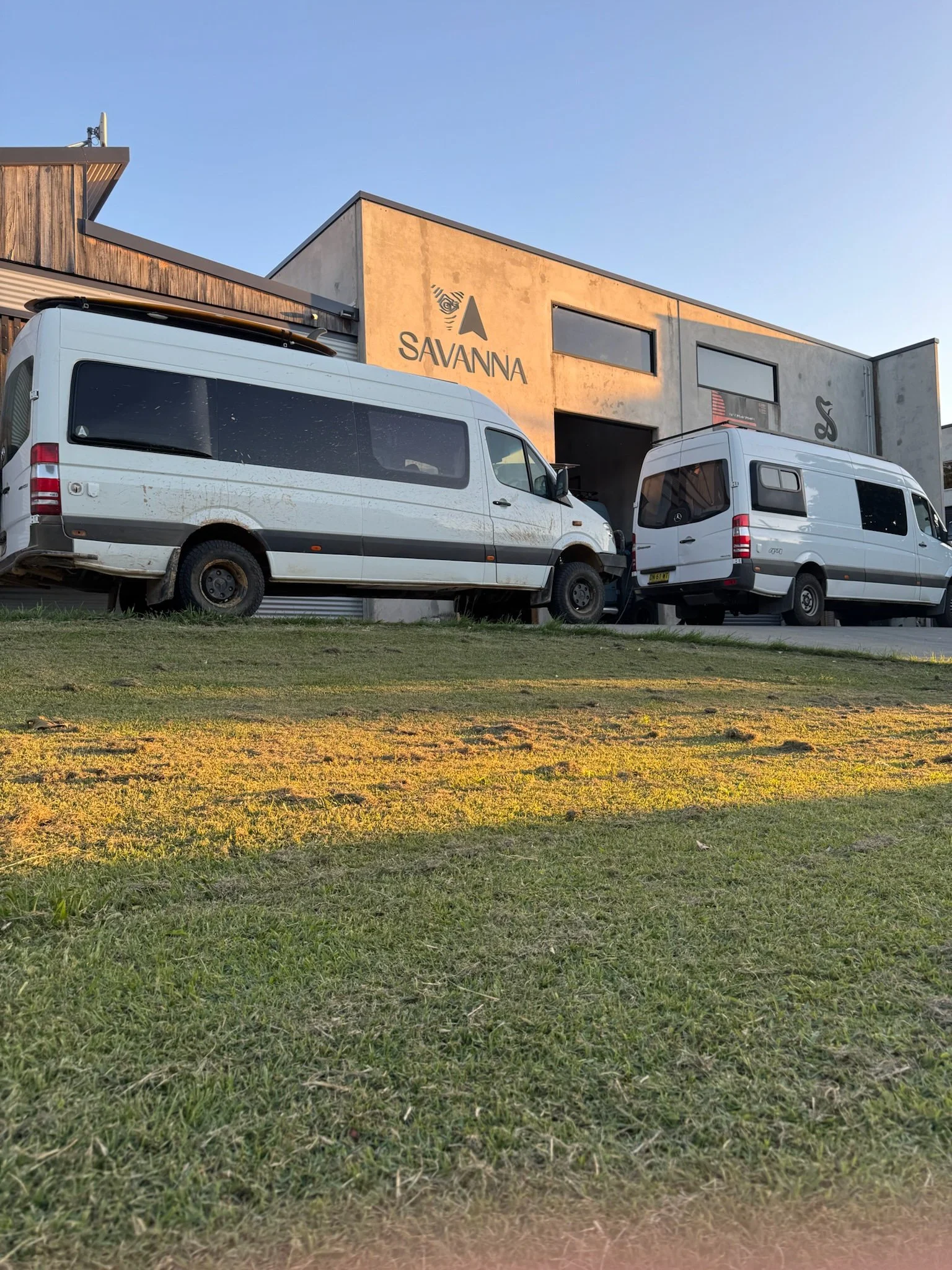 Two white vans parked in front of a building with the sign 'SAVANNA' on it. The building has a modern industrial design with concrete and wooden elements. The scene is captured during sunset with clear skies.