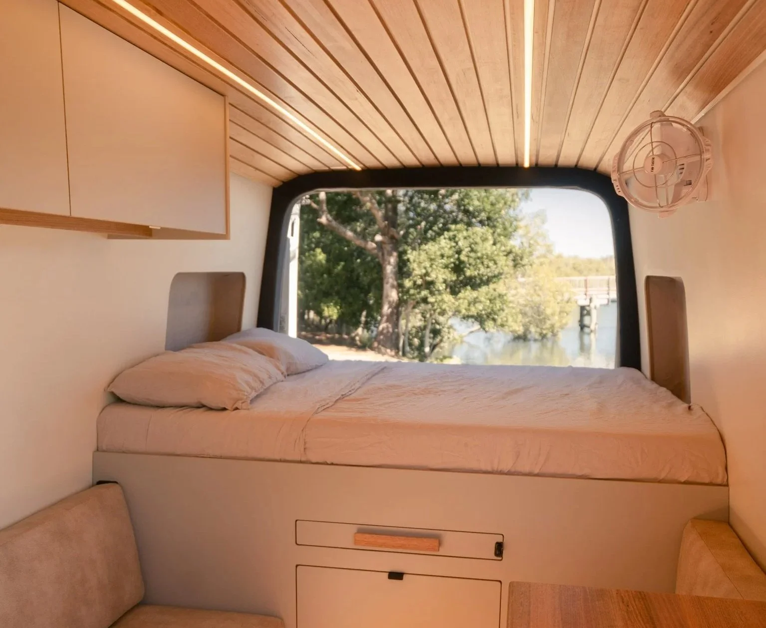 Interior of a modern camper with a bed against the window, a view of trees and water outside, a mounted fan, and wooden panel ceiling.