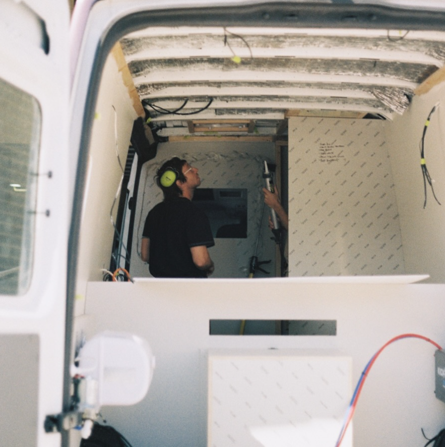 Person wearing safety headphones using a hammer inside a small, partially constructed room or vehicle interior.